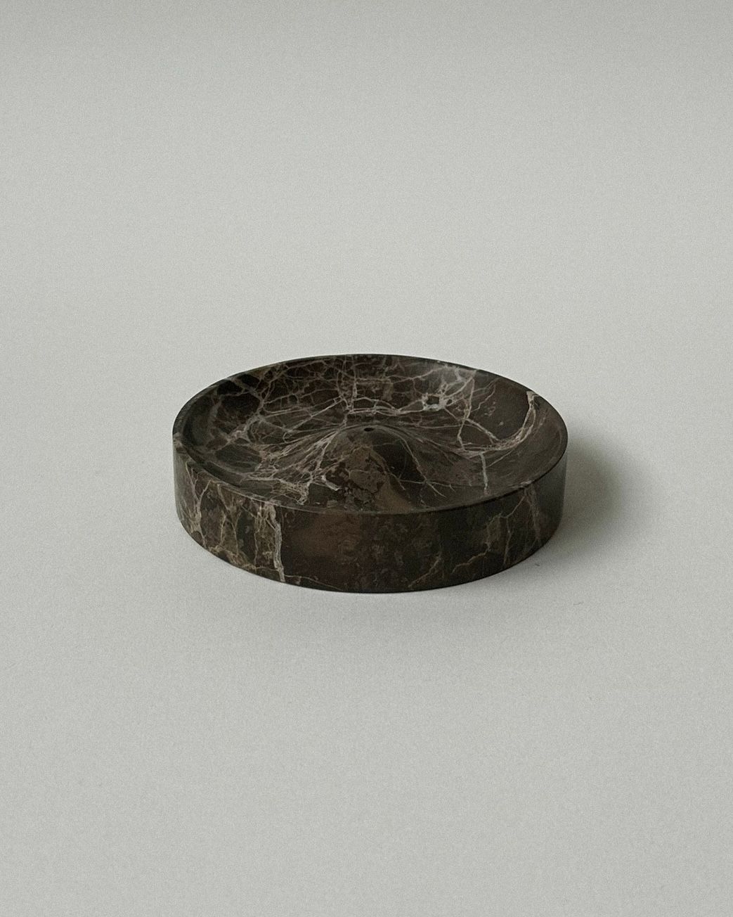 The 'ALTAR' Incense Holder by CARRARA Studio is a round, dark marble tray with white veining, handcrafted from solid stone. It features a low edge and polished finish, shown here on a plain, light gray surface.