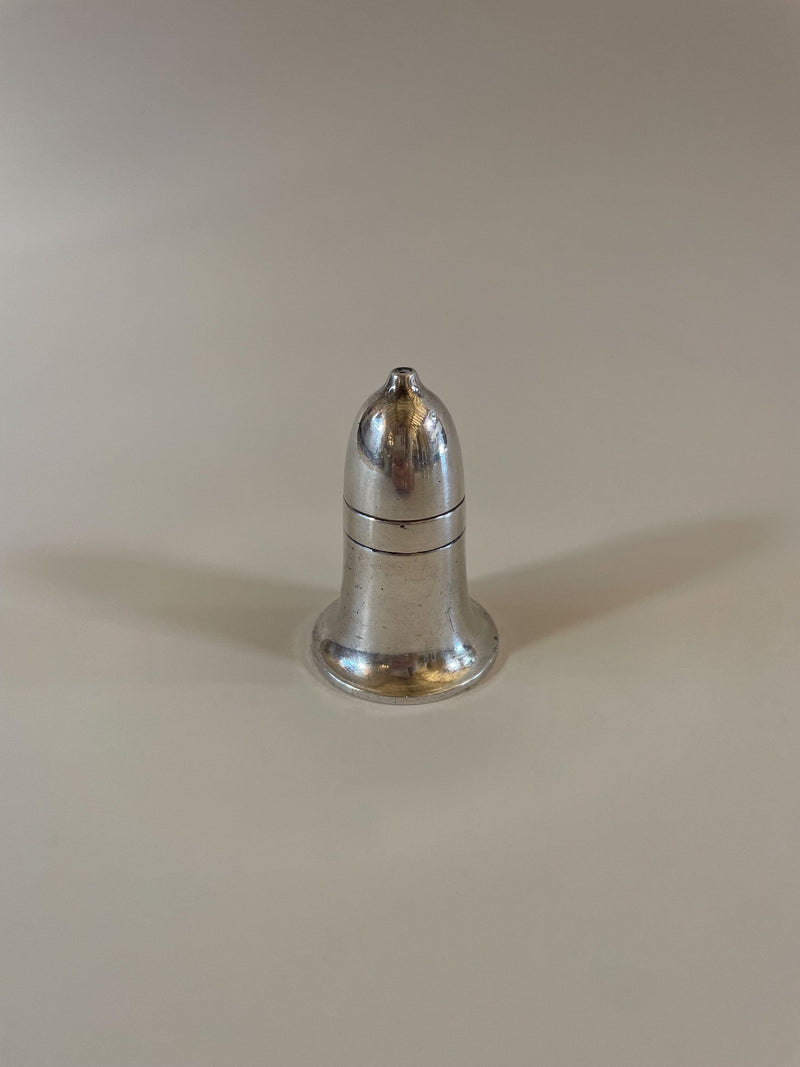 A small metallic bell-shaped object, reminiscent of a Vintage Mappin and Webb Cruet Set by Maud Vaughan, sits on a smooth, light-colored surface. The shiny finish casts a faint shadow to the left against the plain, neutral background.