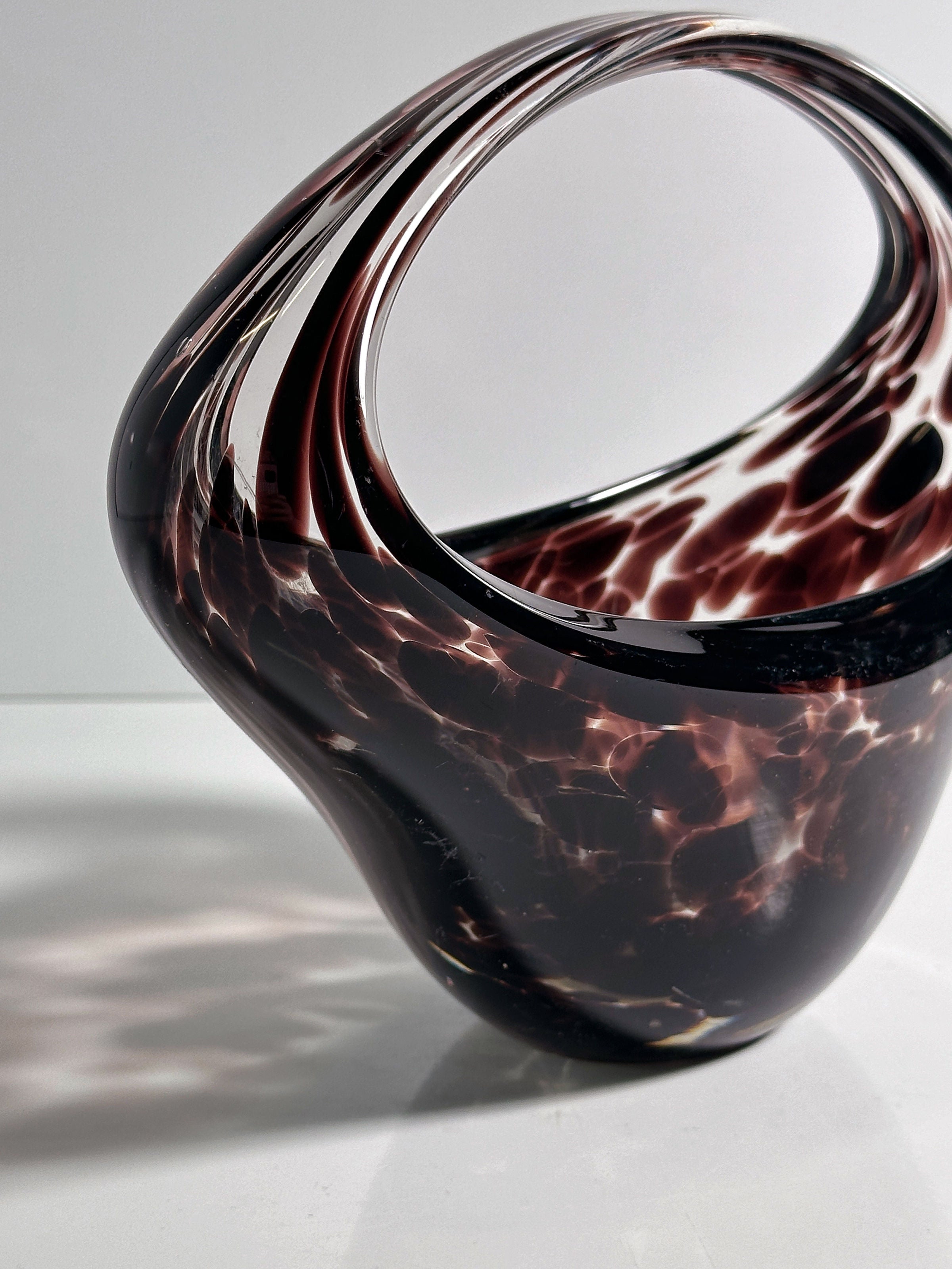 The Murano Tortoise Glass Handle Bowl by Dodo Vintage features a dark marbled pattern reminiscent of a 1970s tortoise look, with a wide, curved base that narrows into an open loop. Its polished surface reflects light beautifully, capturing the dynamic charm intended.