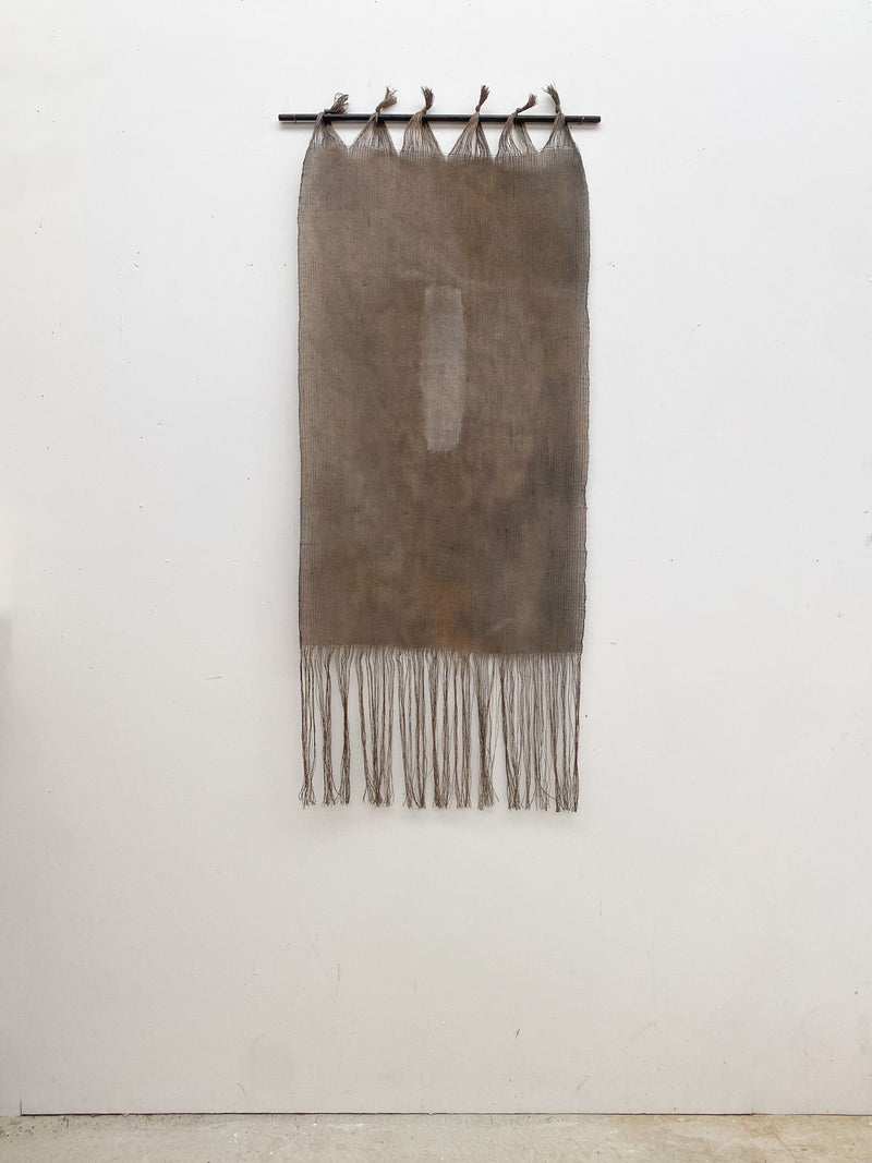 The Moontide tapestry by Sundance Studio hangs vertically from a black rod, showcasing Yuraygir National Park's beauty. Featuring fringes and a central rectangular patch secured by five loops, it stands out against the white wall like Yaegl Country's vast landscapes.