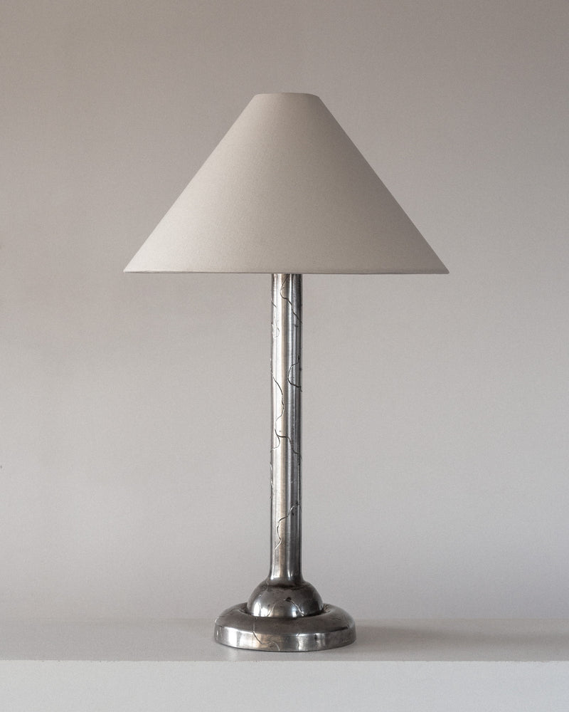 The Spigel Art Deco Pewter Lamp, inspired by 1920s Swedish design, features a pewter base and sleek stem. It comes with a conical beige shade that reflects 1920s elegance, set against a minimalist light background.