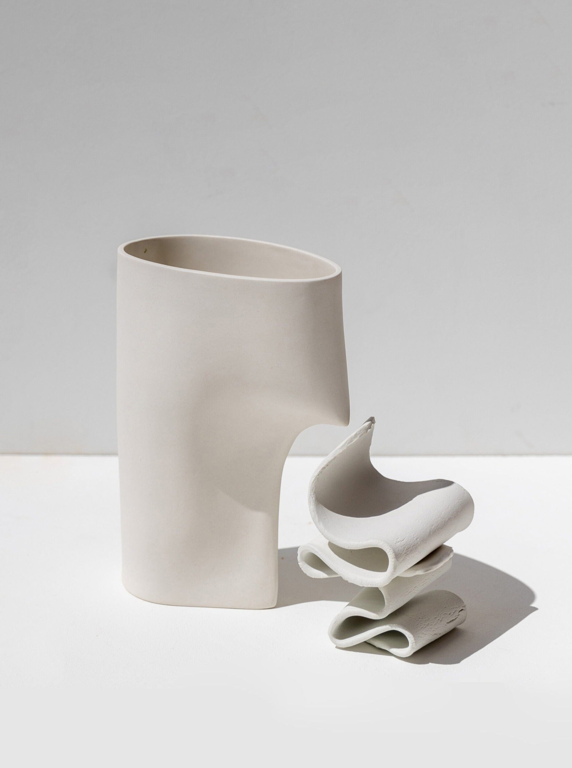 The Draped Vase Apparel by Lisa Allegra is a minimalistic, abstract ceramic sculpture that elegantly stands as a sculptural object. Resembling a tall, hollow rectangle with a curved cutout, this piece is crafted from white porcelain and accompanied by three smaller pieces with undulating, organic shapes. The plain and neutral background highlights the work's smooth surfaces.