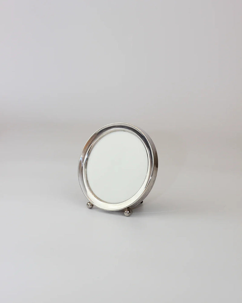 The Round Art Deco Mirror by Boga Avante Shop features a minimalist silver frame and portrait holder, set against a plain gray backdrop. The small, circular tabletop mirror reflects surroundings elegantly with its simple stand.