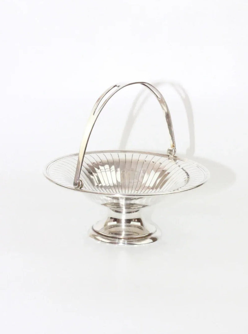 A Silver Fruit Bowl by Boga Avante Shop features a shiny nickel silver round, shallow bowl that sits on a short pedestal base. This vintage piece includes an arched handle and boasts an inner surface with vertical lines radiating outward in a decorative pattern. It is showcased against a plain white background.