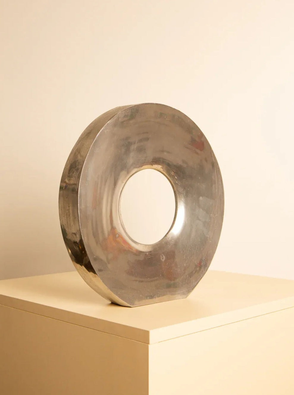 A modern, circular metallic sculpture with a hollow center rests on a minimalist beige pedestal against a light cream background. This contemporary doughnut-shaped piece exemplifies Italian craftsmanship, its reflective surface catching ambient light to create subtle reflections. The "Donut" Vase in Aluminium 80's by Treaptyque perfectly embodies this aesthetic.