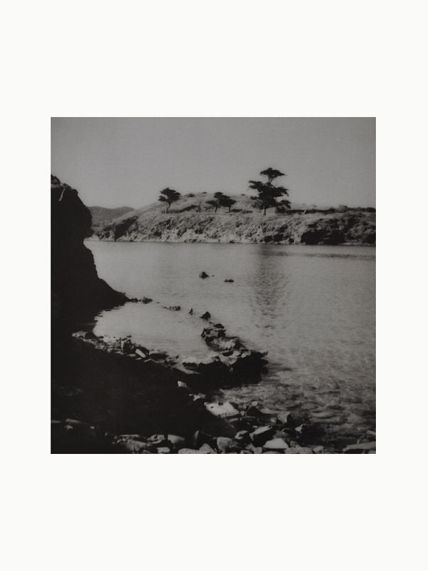 A black-and-white photograph of a serene body of water bordered by rocky terrain and sparse vegetation. The distant shoreline features a few isolated trees and rolling hills under a clear sky, reminiscent of the captivating scenes in Maison Plage's photography book, Valérie Prot: Cadaqués.