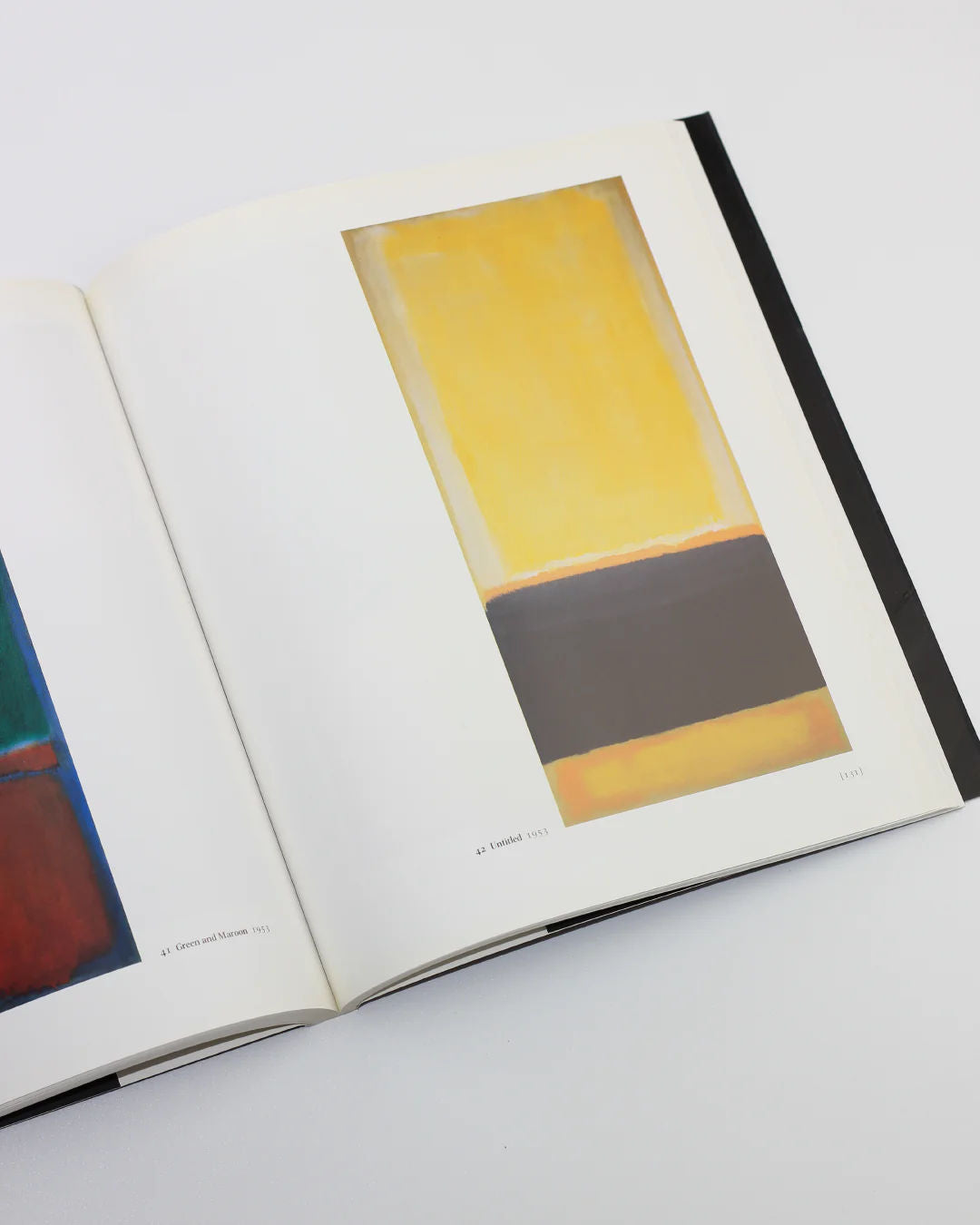 The Mark Rothko Book by Boga Avante Shop opens to an Abstract Expressionism piece showcasing large vertical color blocks. A prominent yellow section tops the art, complemented by a thick black band beneath, all set against a light gray backdrop.