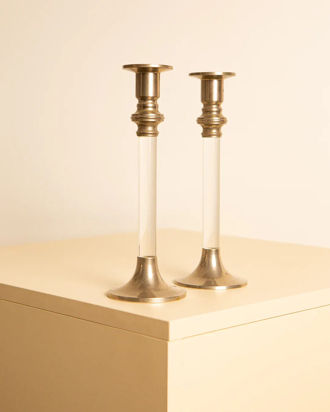 Two Treaptyque Pair of 80's silver and lucite candleholders stand on a cream-colored surface against a matching cream background. The slender candleholders, reminiscent of retro '80s candleholders, feature silver bases and tops, seamlessly combining classical and modern design elements.