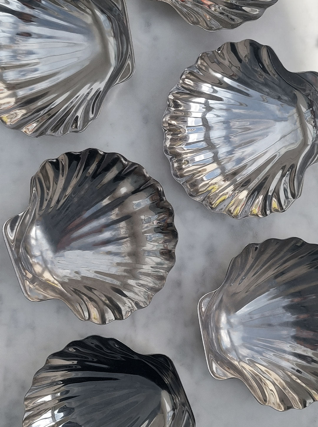 A collection of shiny, stainless steel Set of 6 Small Shell Dishes by Les Objoies arranged on a light grey marble surface. The molds have a reflective surface, capturing various light reflections and angles, perfect for elevating your dining experience with elegance and charm.