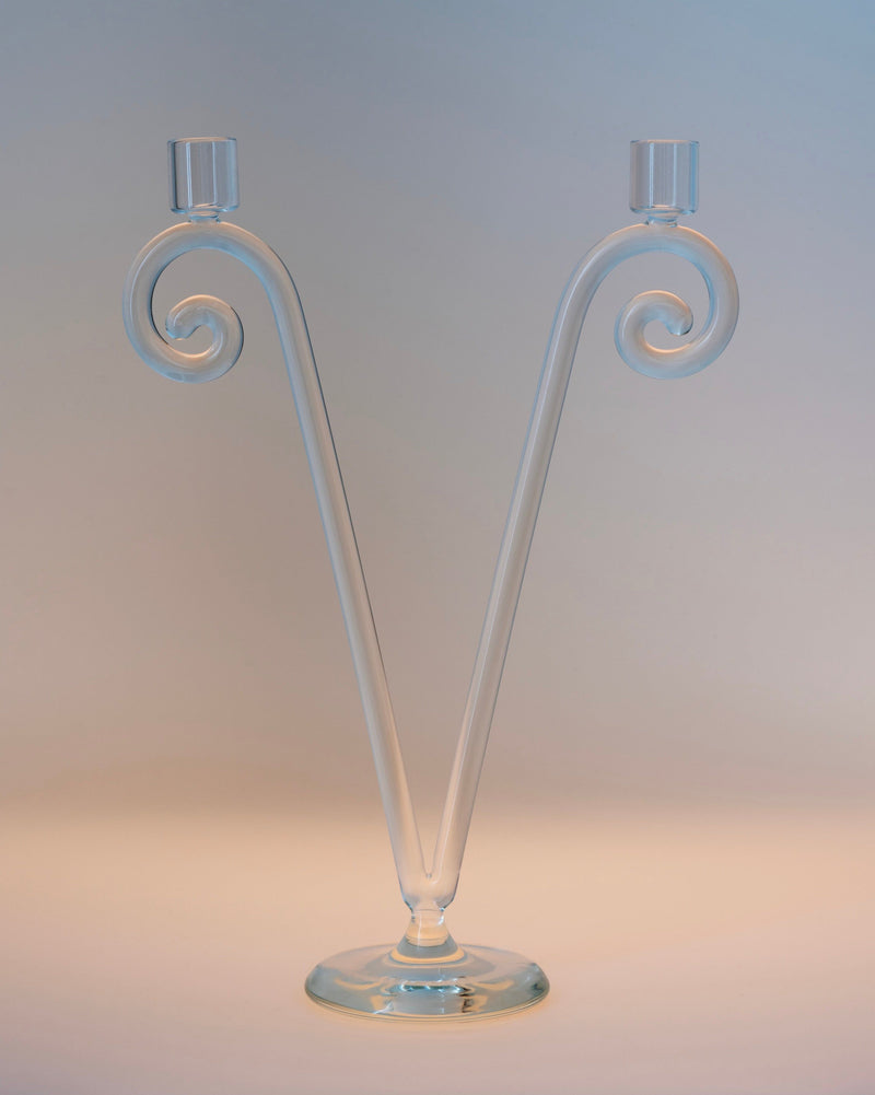The Double Stem Spiral Candleholder by Sophie Lou Jacobsen features two clear glass arms curving upward from a central base in a V shape, each ending in a candle socket. Set against a softly lit, plain background—ideal for adding vintage dining charm.
