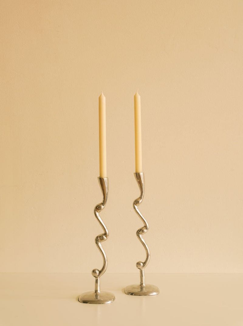 Two tall, slender candles in elegant, Les Objets Amsterdam Curly Candleholders stand against a plain, light beige background.