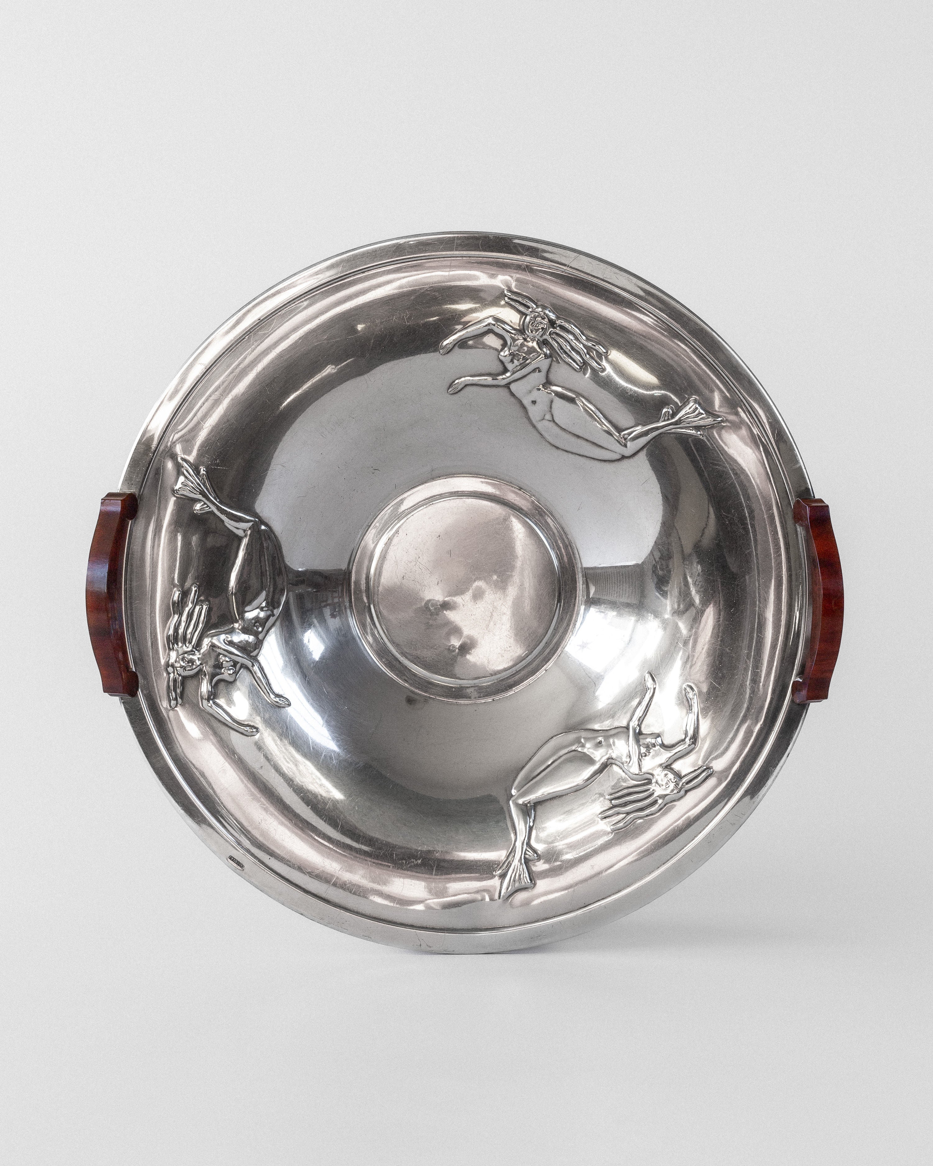 The Spigel Decorative Silver-Plated Dish from 1930s Sweden features a round Art Deco design with two dark brown Bakelite handles and three evenly spaced decorative figures of leaping gazelles on a plain light gray background.