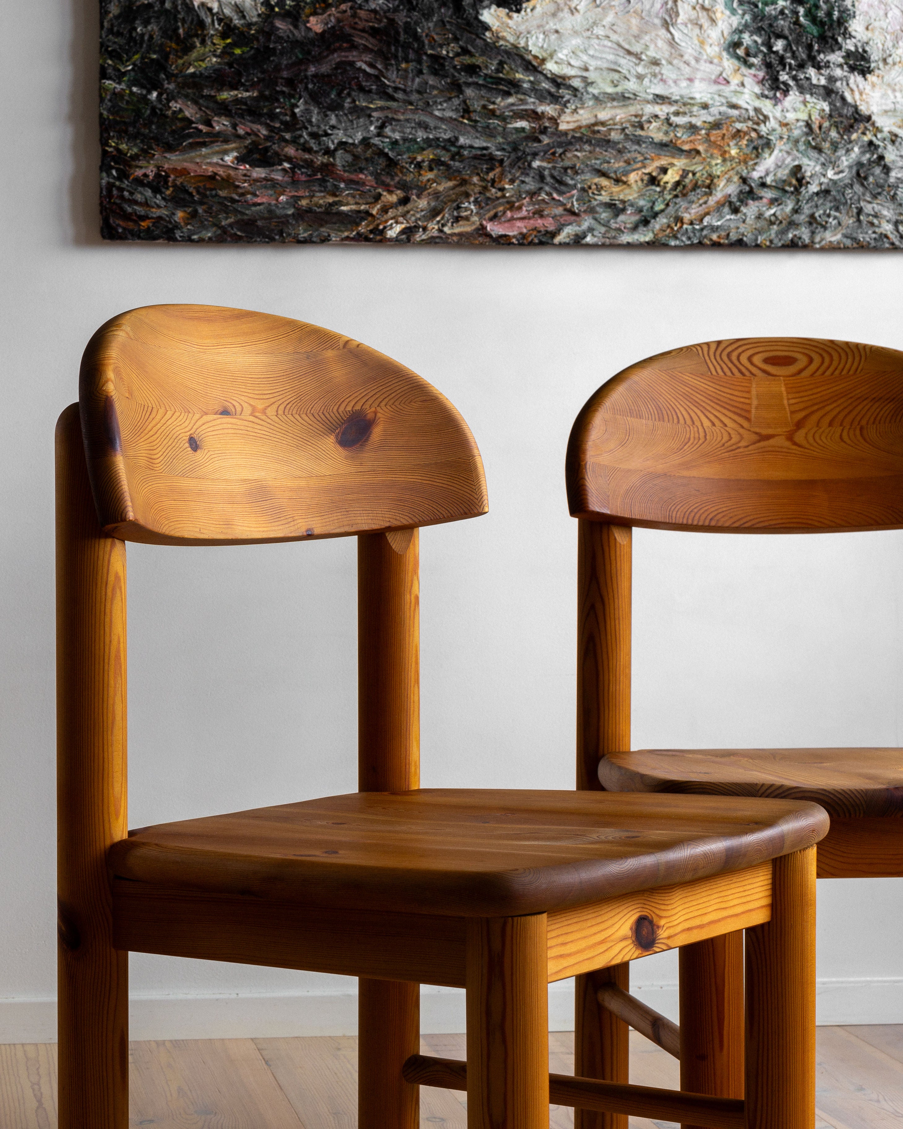 Two of the 4 Pine Chairs by Rainer Daumiller, created in Denmark in the 1970s and offered by Spigel, are positioned in front of a textured, abstract painting mounted on a white wall. These solid pine furniture pieces have a natural finish that showcases the wood grain and are placed on a wooden floor. The scene is lit with soft, natural light.