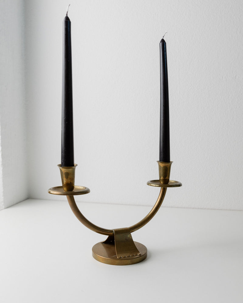 The Bottega Jacobs Art Deco Candleholder from 1960s Italy, made of brass, holds two unlit black candles. It sits gracefully on a white surface with a minimalist design and curved base against a plain light-colored wall.