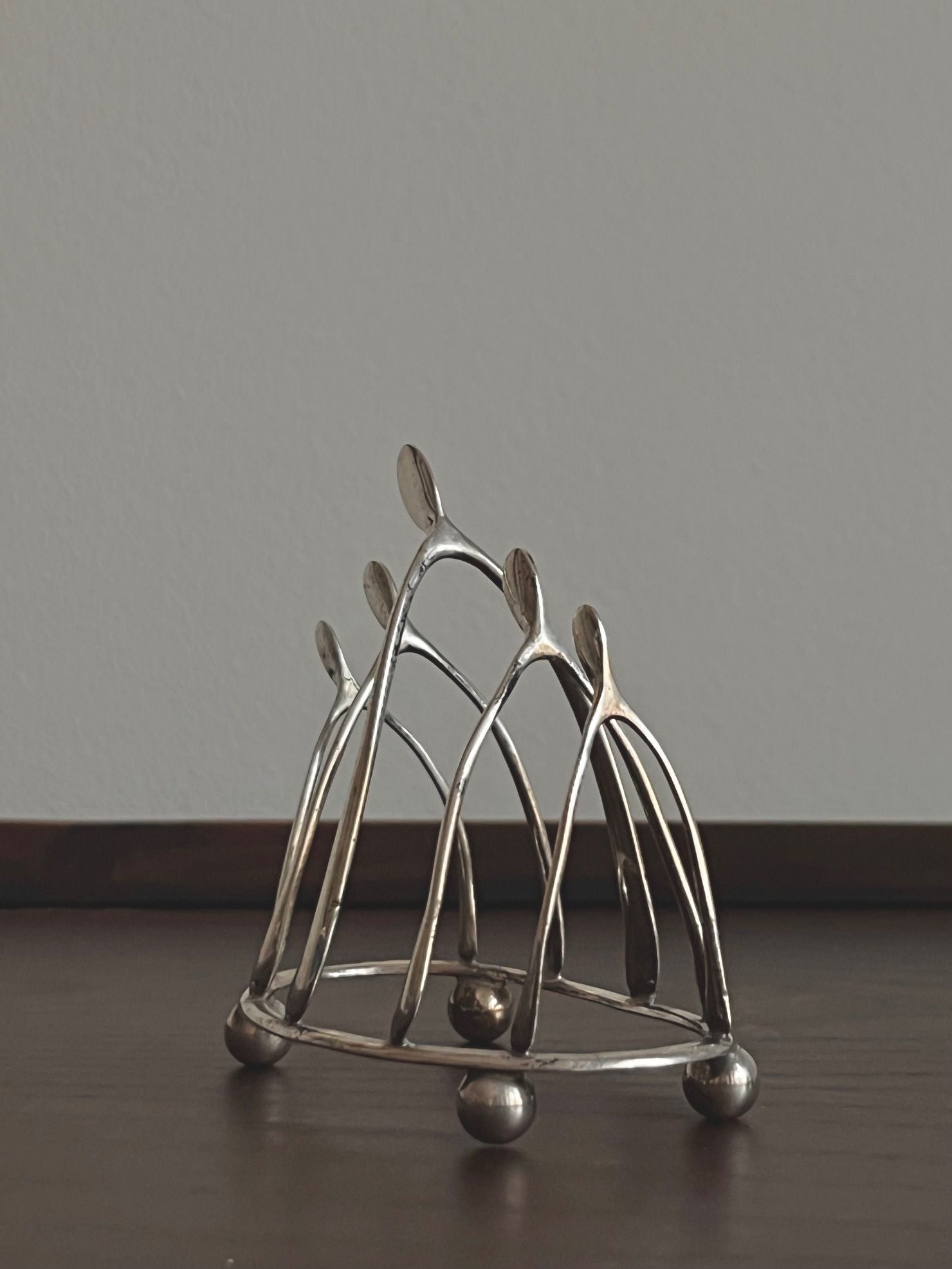 The Wishbone Silver Toastrack by Médecine features abstract human figures with elongated limbs standing in a circle on a wooden surface. The silver metallic design evokes unity and balance as the figures elegantly hold hands, reminiscent of an antique toastrack.