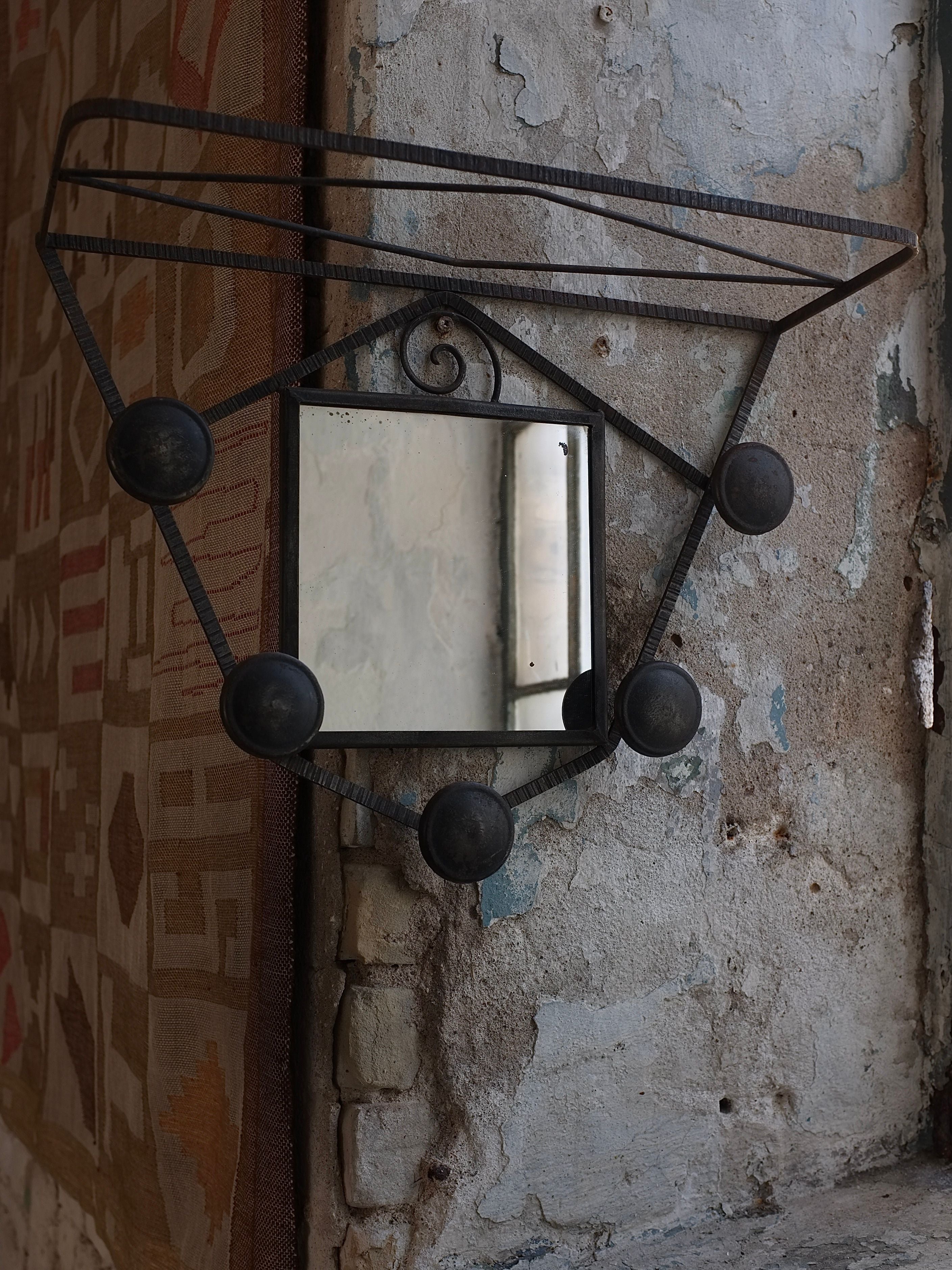 A small, square mirror with a decorative metal frame from the 1930s, known as the Art Deco Wrought Iron Wall Mirror Coat Rack by Veter Vintage, hangs on a textured, weathered wall. The frame features circular accents and metal rods creating a geometric pattern. The worn wall has peeling paint and exposed brick, with part of a patterned rug visible on the left.