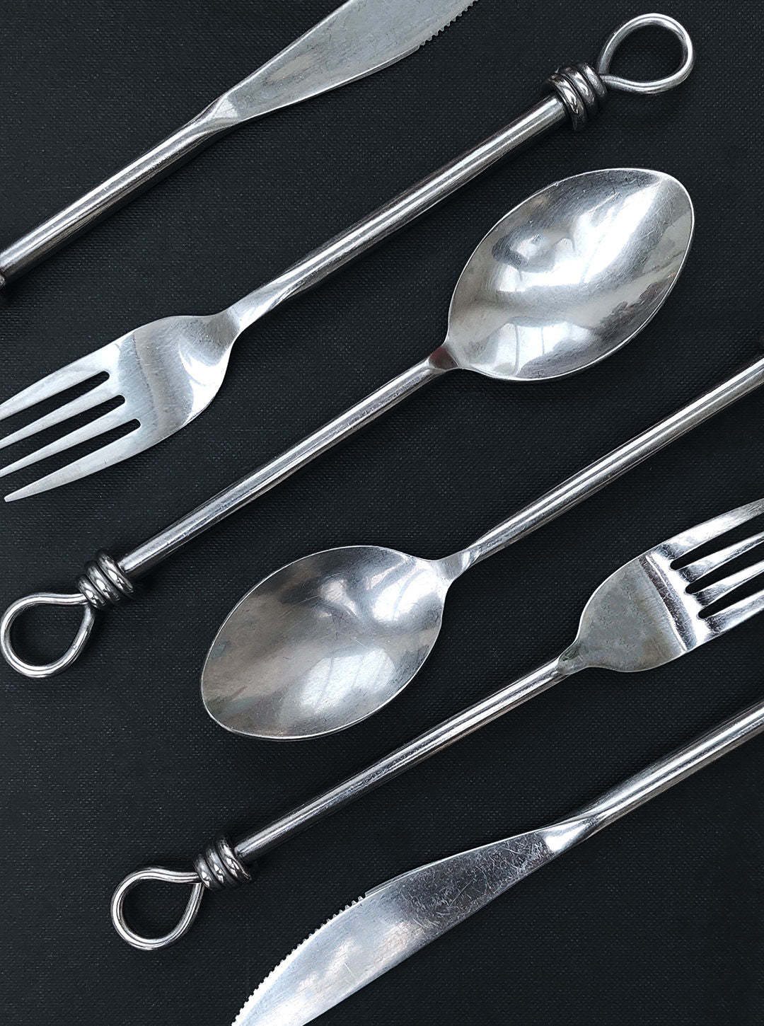 A Set of 6 Cutlery from the brand Les Objoies is arranged diagonally on a dark background, showcasing each piece's decorative loop handle design, which adds an elegant touch to the collection.