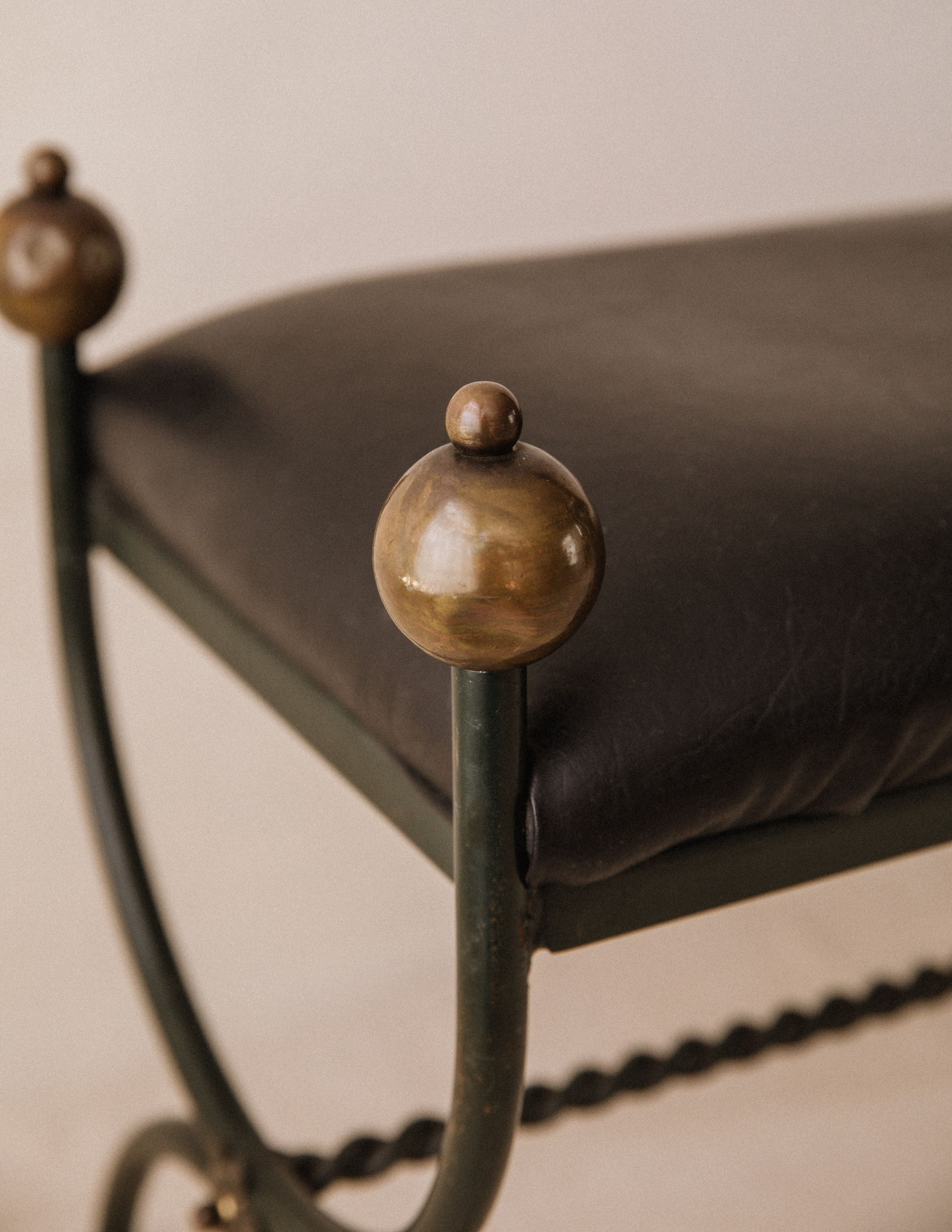 Close-up of the Mid-Century Wrought Iron Bench by Arcā Collections, featuring a dark leather seat, spherical brass finials on each corner, and twisted metal bar accents. The neutral background keeps the focus on this Jean Royère-inspired design.