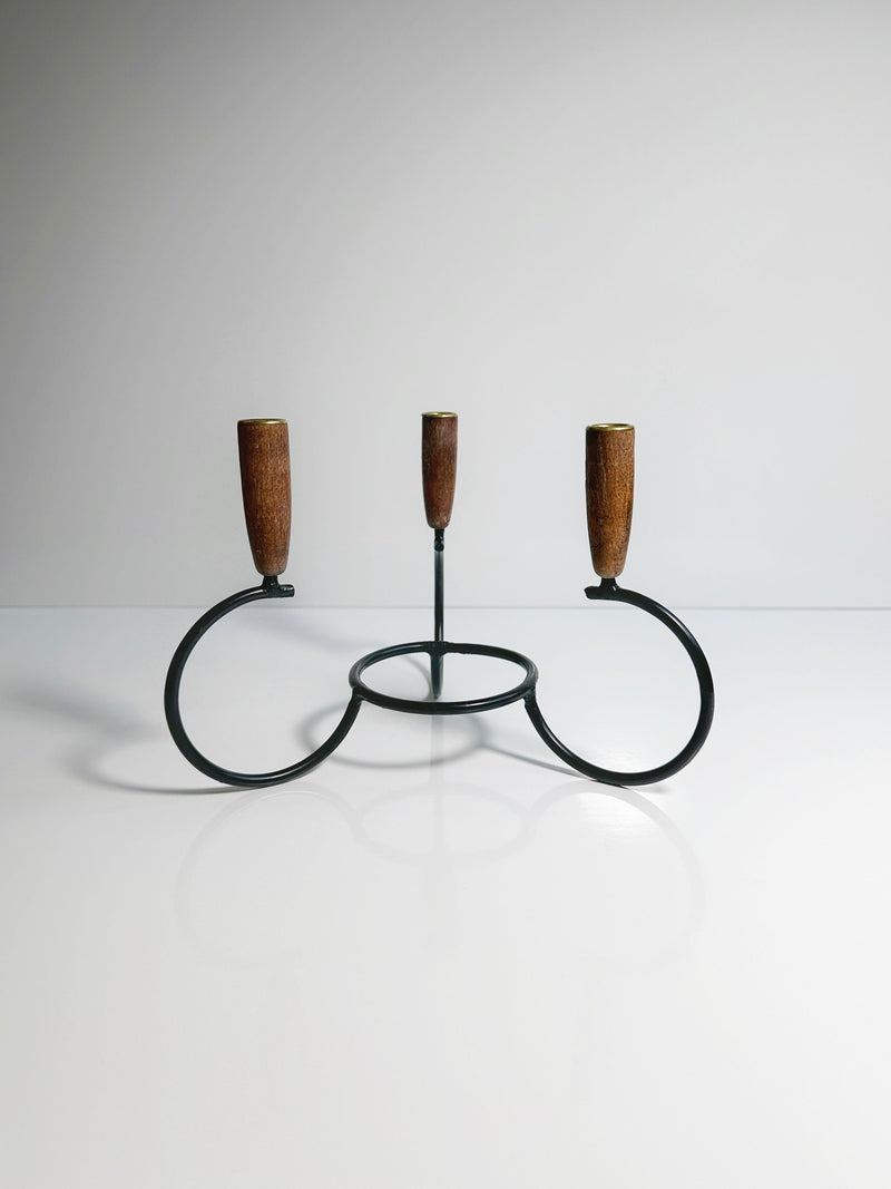 The Danish Candleholder 1960s by Dodo Vintage is a minimalist black metal candle holder that features three circular bases, each topped with teak ends. Designed to hold three candles, it is set against a plain, light background to create a clean and simple aesthetic.