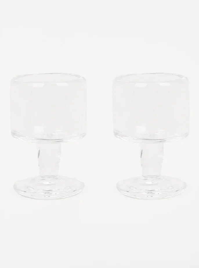 Two clear glass goblets with thick stems and circular bases are placed side by side against a plain white background. These elegant goblets, the 0405 Stem Glass Set of 2 | Clear | Small, hand-blown in Europe by FRAMA, boast a simple yet refined design.