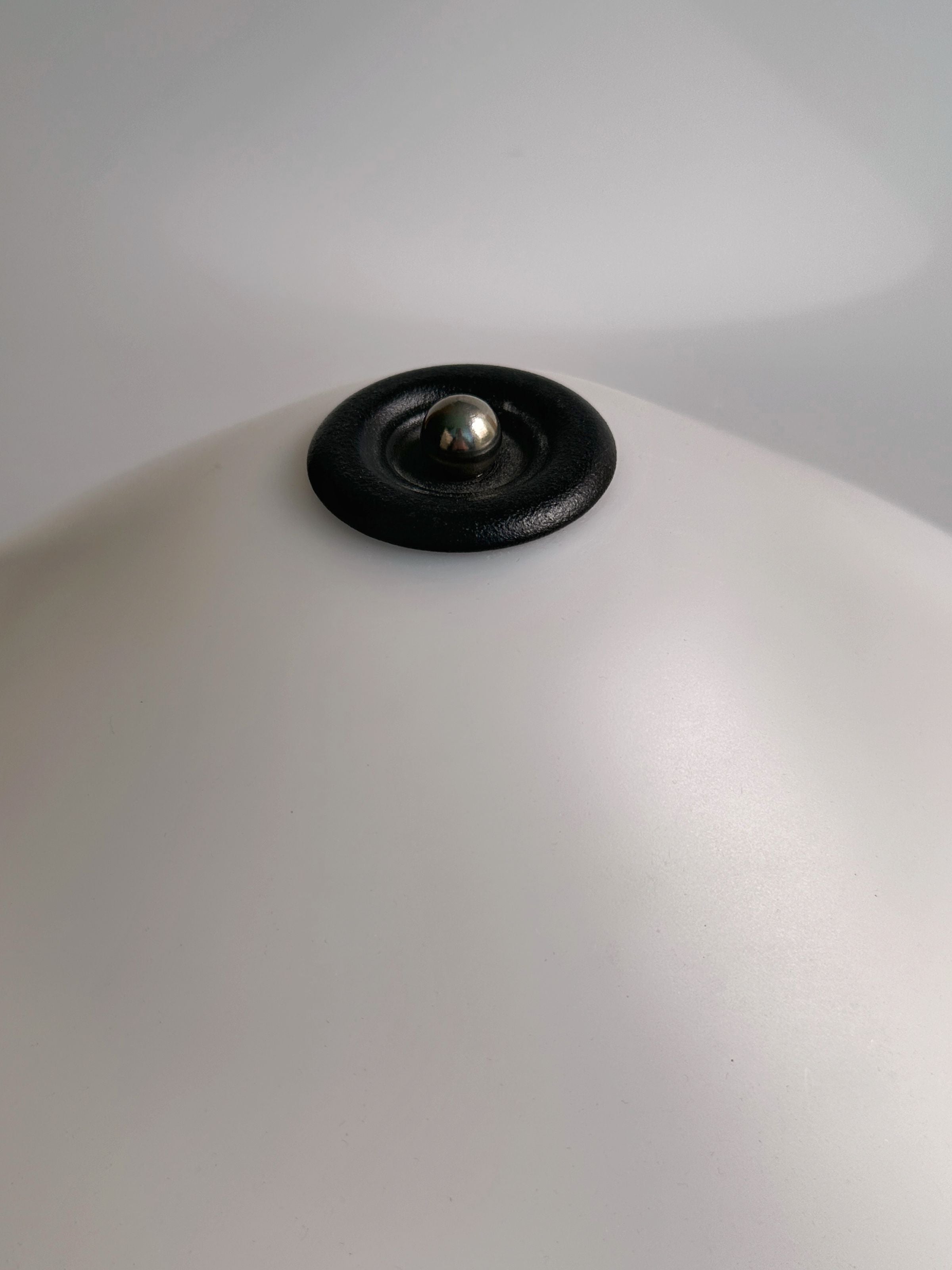 A close-up showcases the 1950s Scandinavian Banker Lamp by Dodo Vintage, featuring a matte white dome shape and a central black lacquered metal detail with a small metallic knob. The softly lit background highlights the lamp's quintessential Scandinavian Mid Century style.