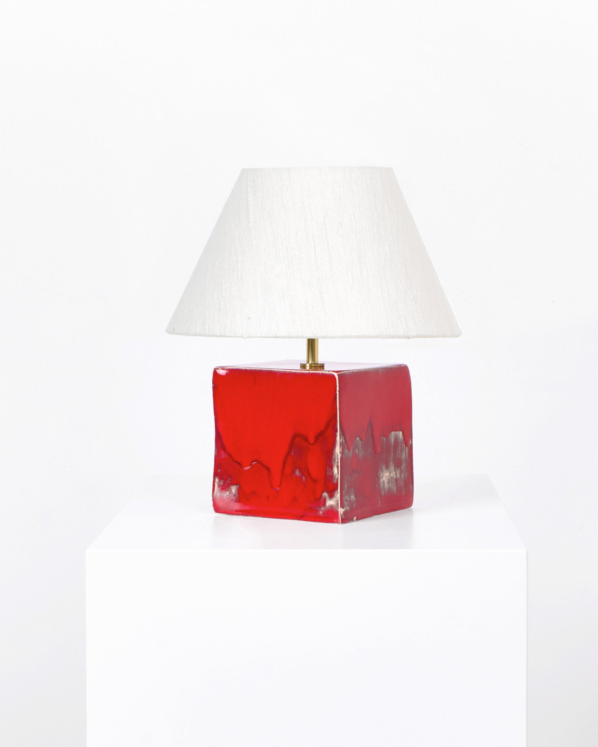 The Arouca Table Light Red by Project 213A features a red, glossy, cube-shaped ceramic base with abstract patterns and a white cone lampshade. This artisanal lamp is displayed on a white pedestal against a plain background.