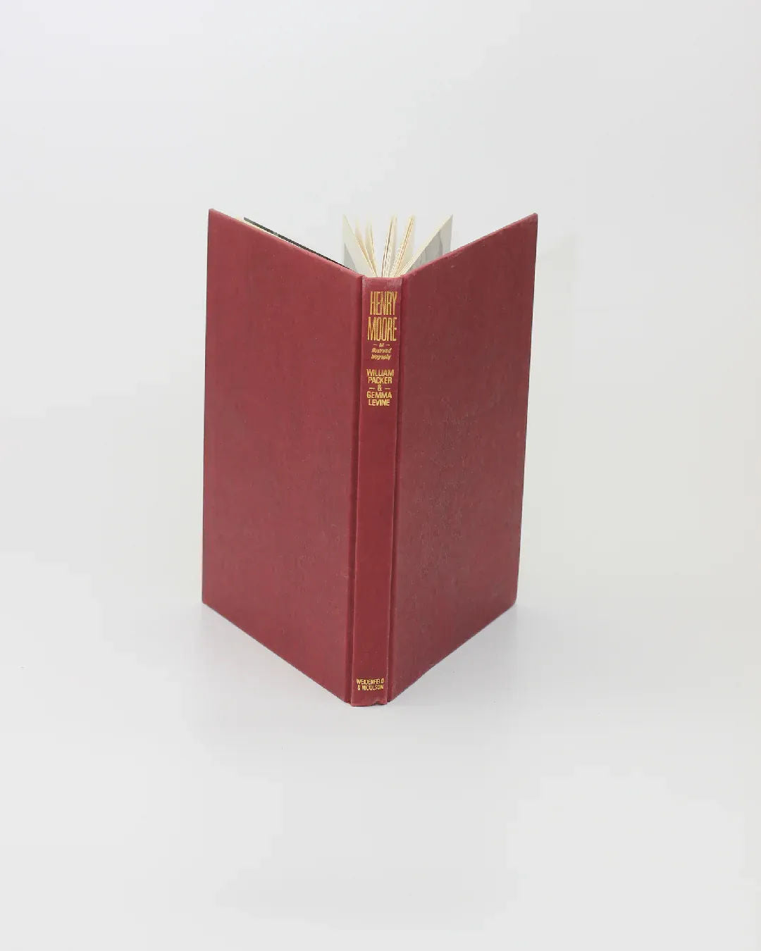 A vintage red libro titled "Henry Moore, An Illustrated Biography" from Boga Avante Shop stands upright on a plain white background, with gold lettering on its spine and slightly open cream-colored pages, reflecting the elegance of escultura moderna.