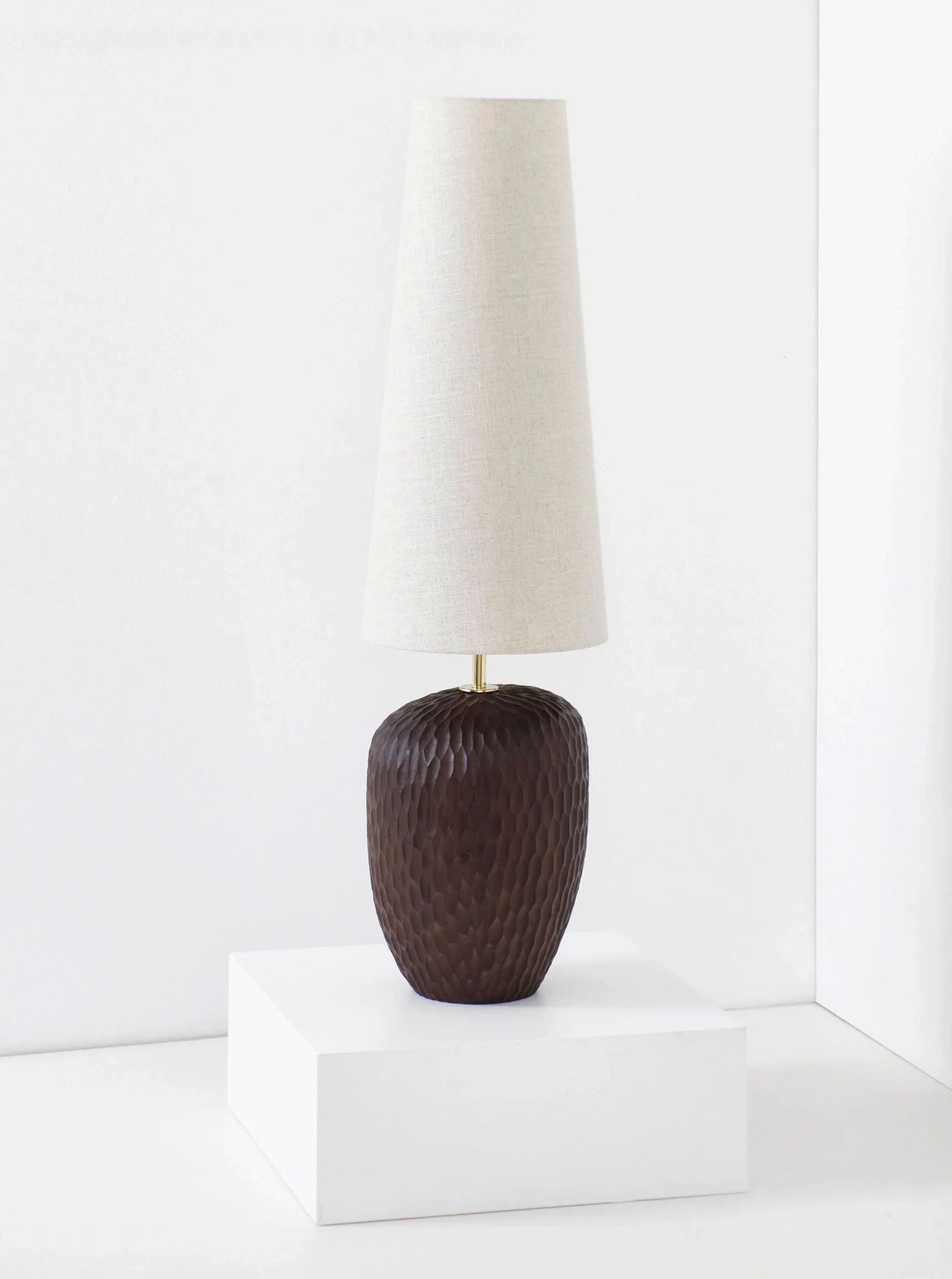 A tall, modern Project 213A Large Foz Lamp with a textured dark brown solid wood base and an elongated, tapered white lampshade is placed on a white pedestal against a plain white background.