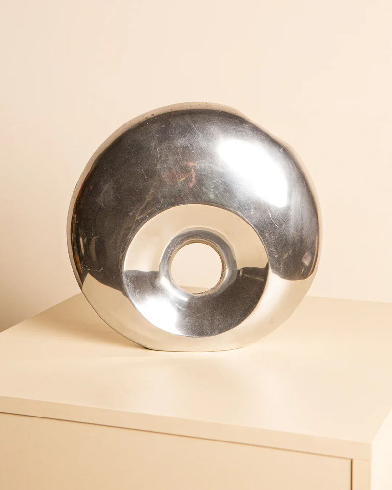 The "Cercle" Vase in Aluminum 80's by Treaptyque, part of a private collection, is a reflective silver donut-shaped sculpture on a light surface with a plain background. Its elegant metallic torus design and smooth finish echo the 1980s aesthetic.