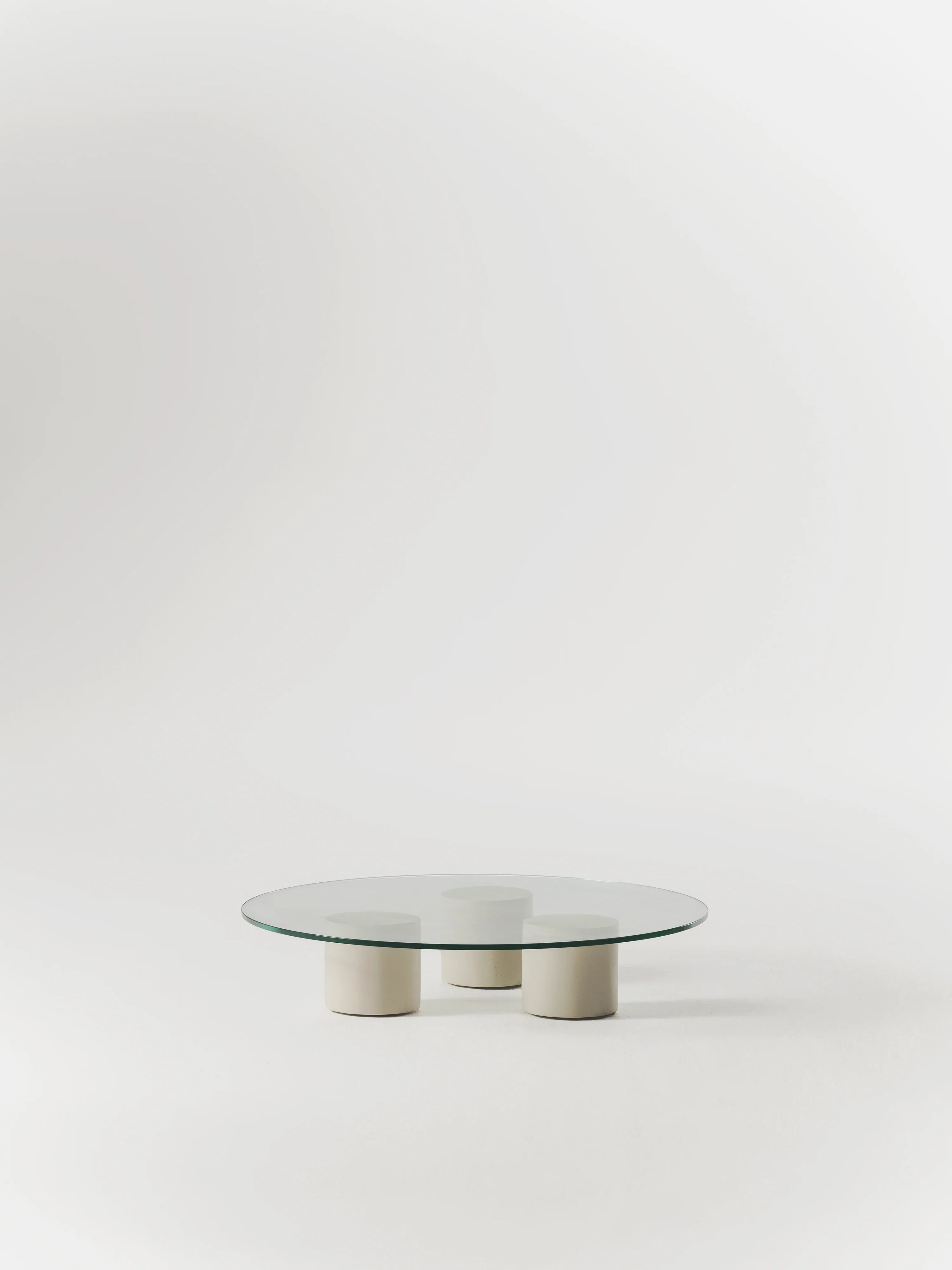 The Carmene Glass Coffee Table by MarlotBaus showcases a round, tempered glass top supported by three cylindrical white bases, set against a plain white background.