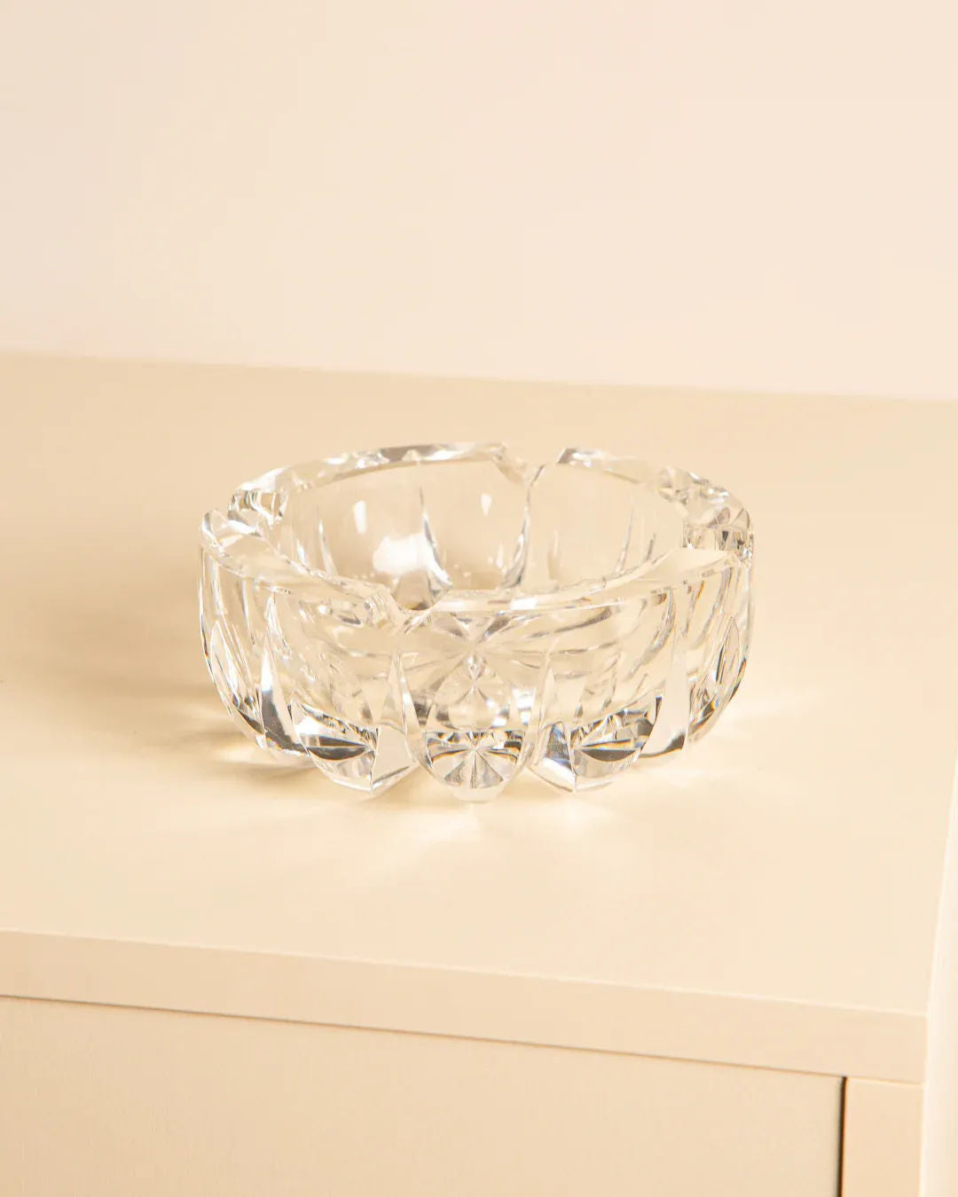 A clear, intricately cut crystal bowl rests on a light surface, evoking a 1970s Saint-Louis "Ambassadeur" D120 ashtray by Treaptyque. Its facets sparkle with reflections and highlights against the plain beige background.