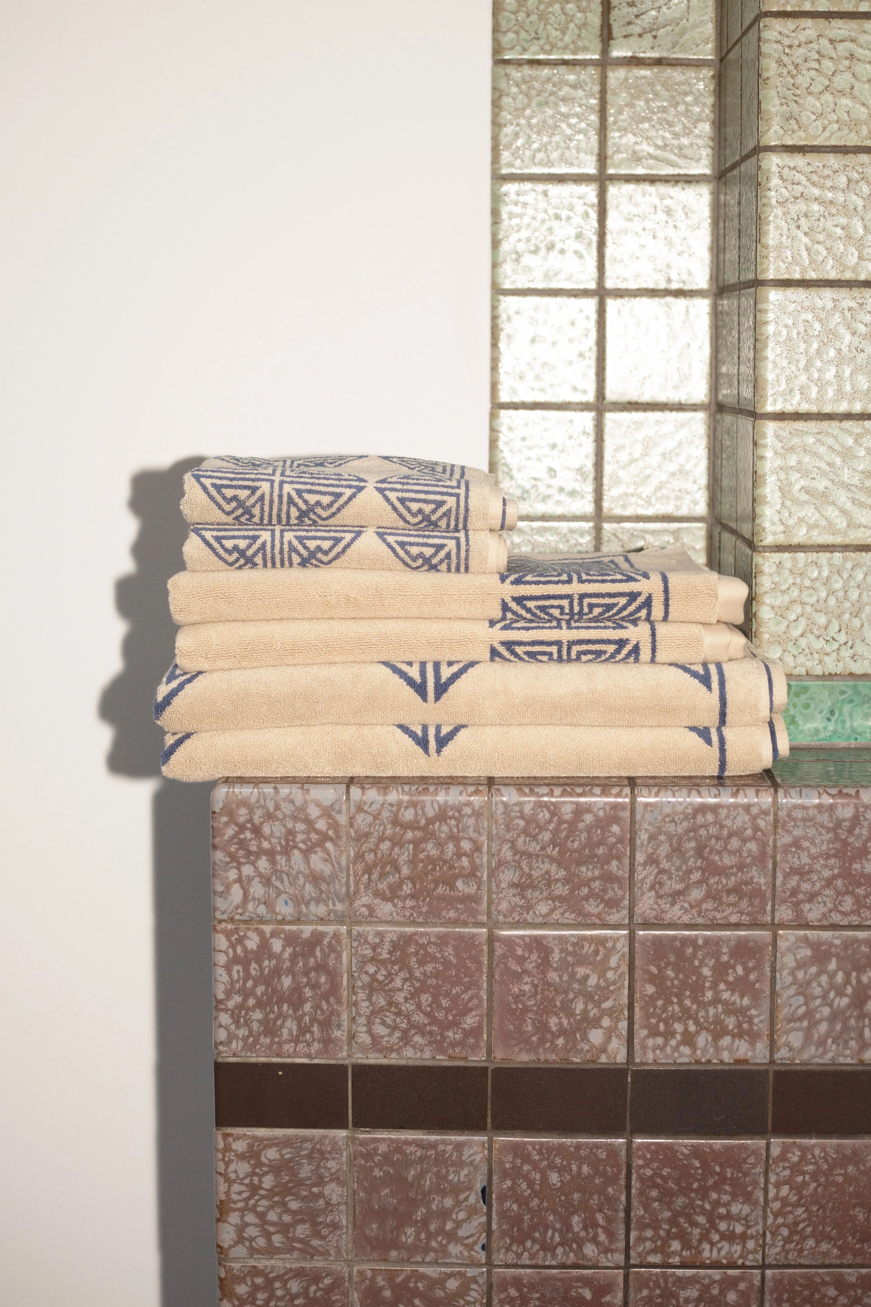 Folded towels with geometric designs stacked on a tiled bathroom shelf. The wall features a mix of plain and textured glass blocks, casting shadows and light patches. Among the items is an Autumn Sonata Agnes Bath Mat made organic materials.