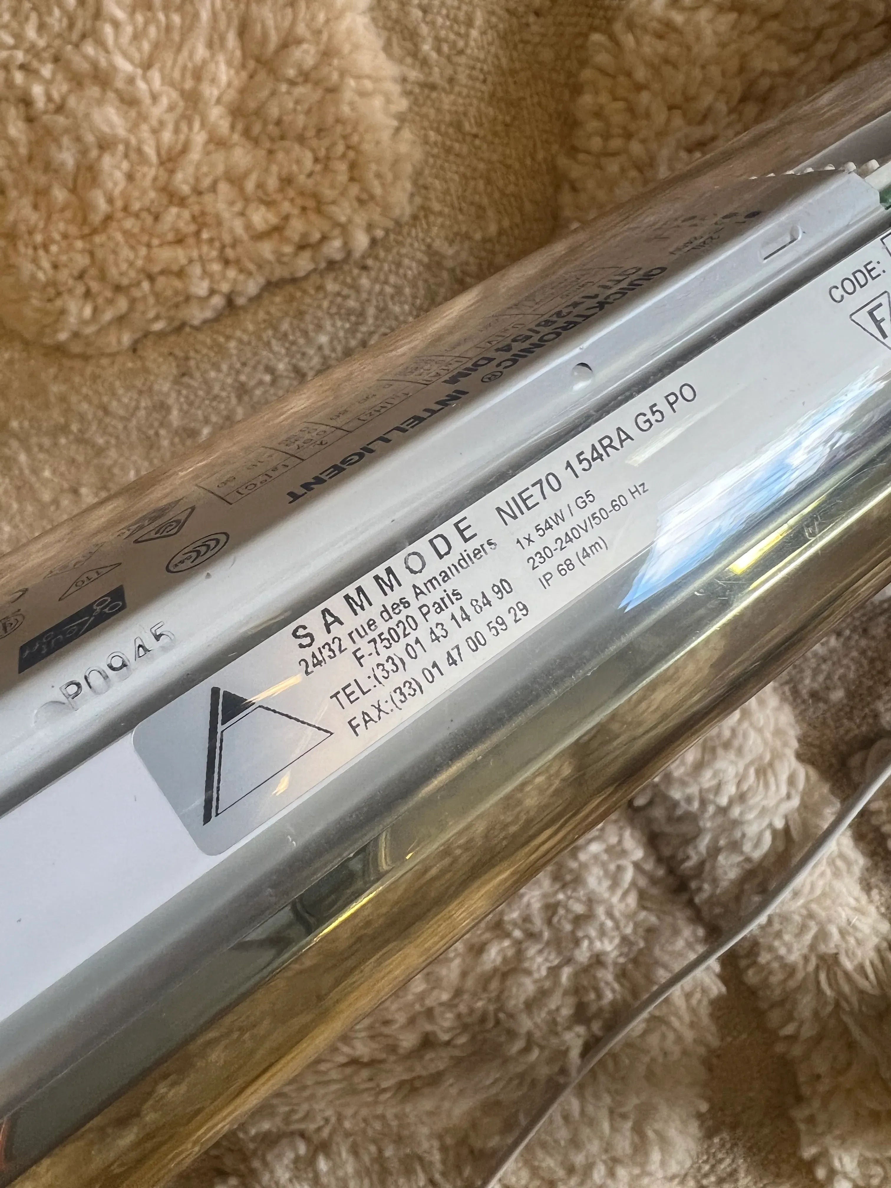 Close-up of a CU-RA-TED Sammode Light Tube, model number N1701548RA G5PO, with an address, contact information, and specifications printed on the side. The elegant French architectural lighting piece is placed on a textured beige surface.