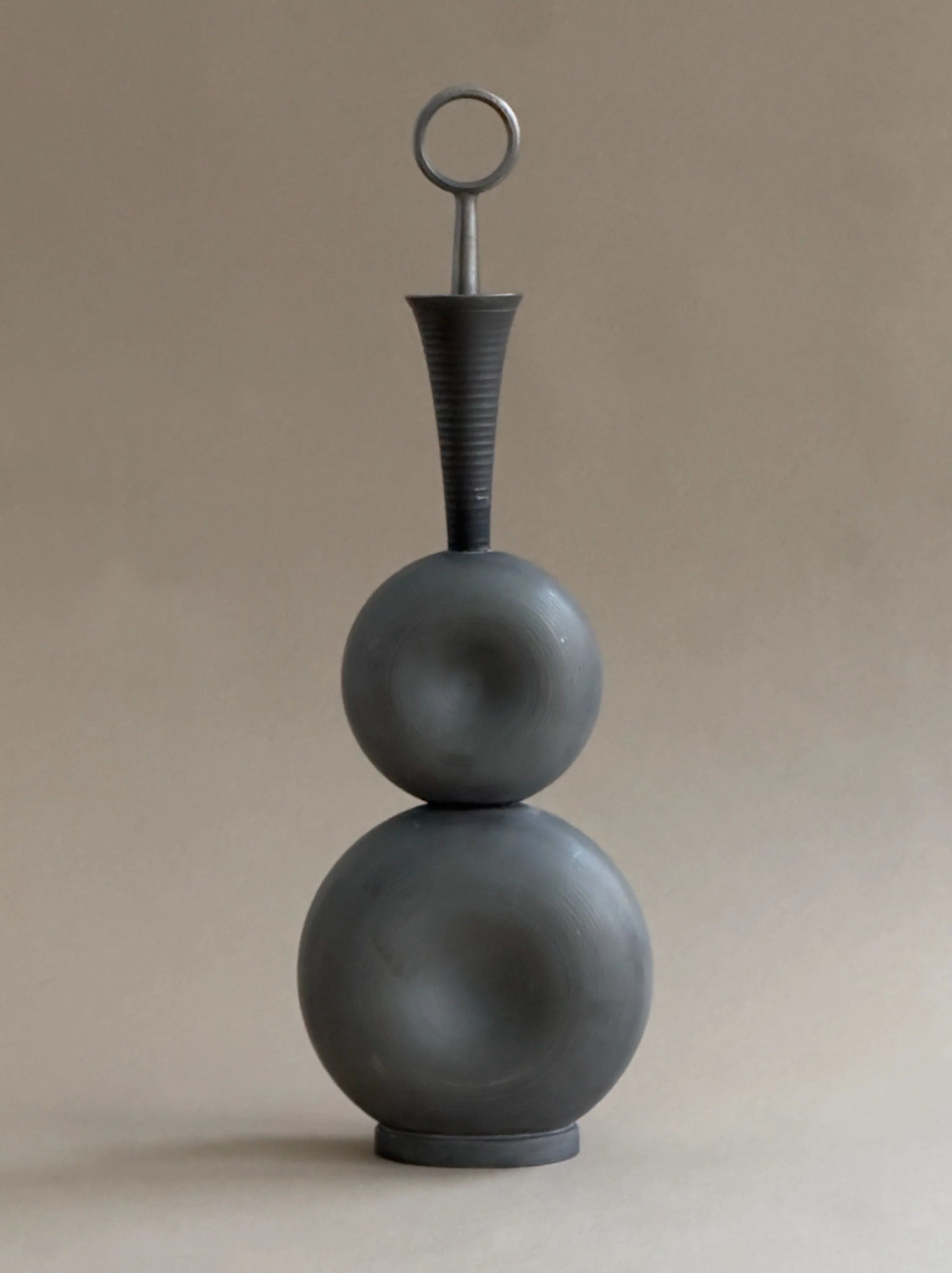 A modern sculpture, akin to a Gunnar Havstad piece, resembles the Galerie Storm's Modernist Pewter Decanter with two stacked gray spheres, a narrow conical top, and circular handle. The neutral background accentuates its minimalist design.