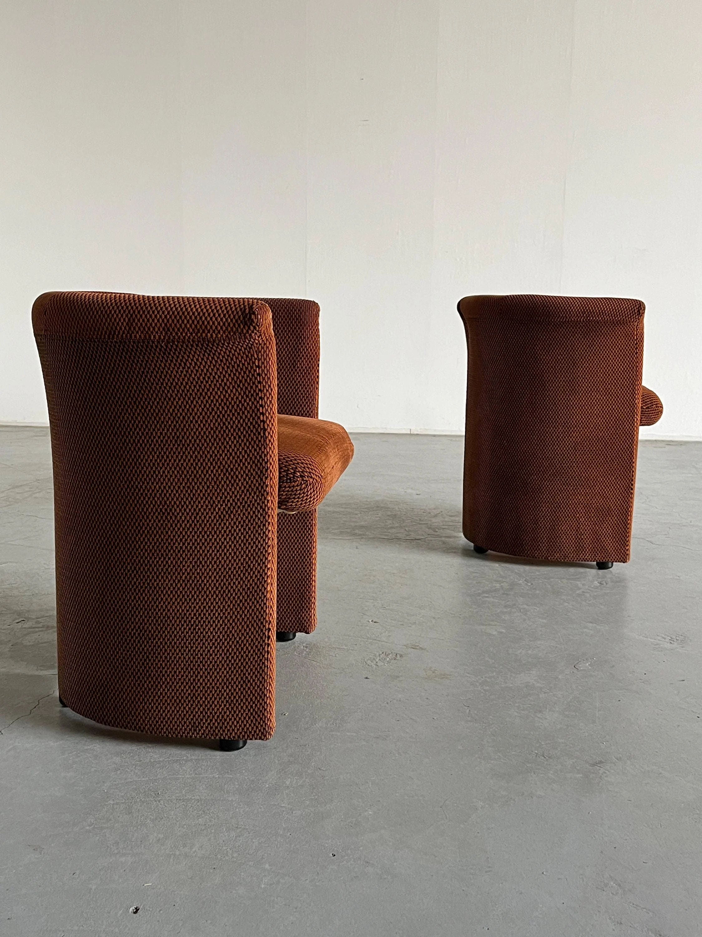 In a minimalist room with plain white walls and a gray floor, the Pair of Postmodern Armchairs by Cherry Cargo are positioned in opposing directions, highlighting their curved backs and textured brown upholstery.