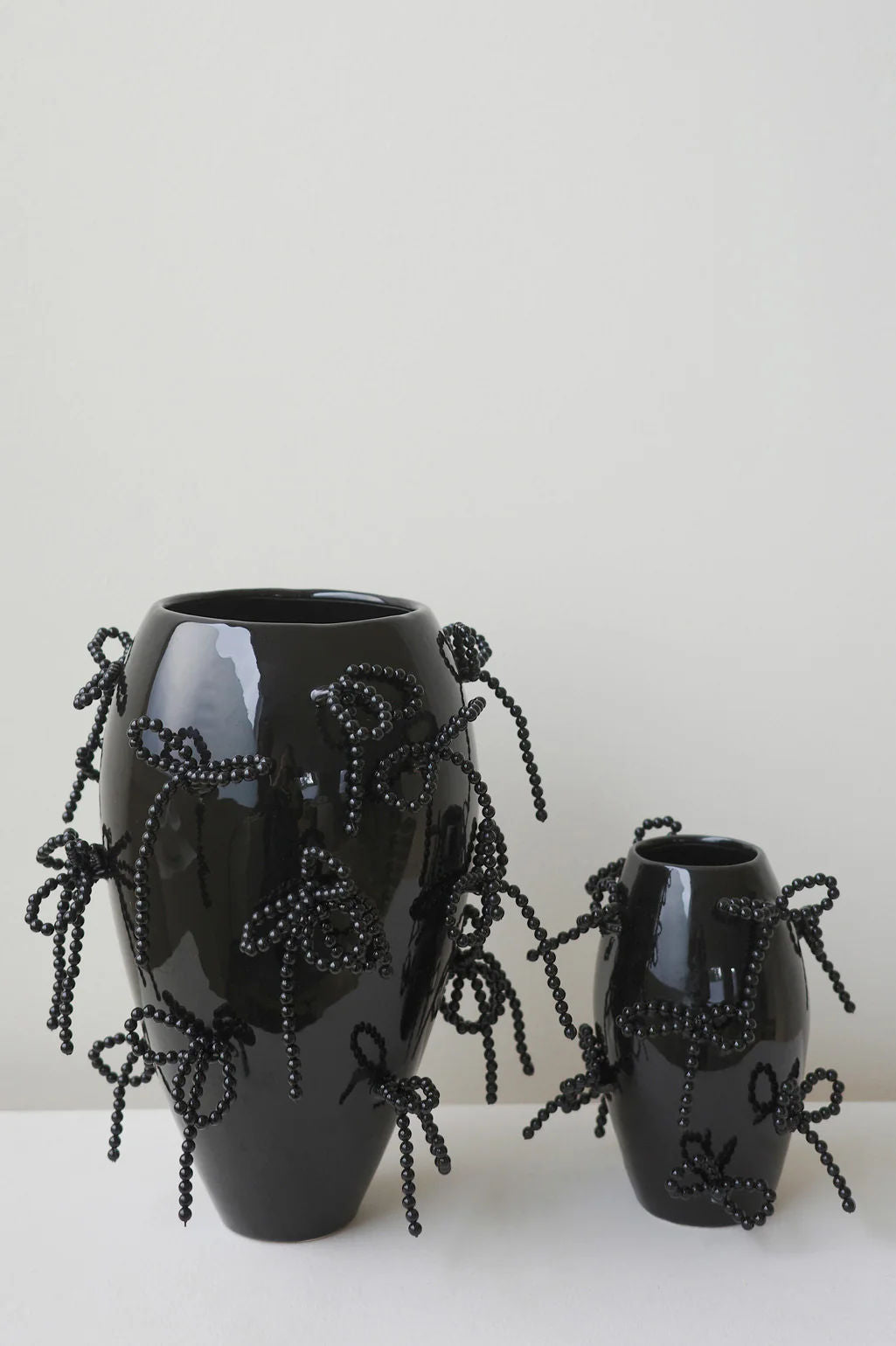 Completedworks' "Pearly Pearl - Large Vase In Gloss Black" features a sculptural design with intricate black beaded bows resembling faux pearls, complemented by a similar smaller vase against a plain white background. Both are handcrafted ceramic pieces.