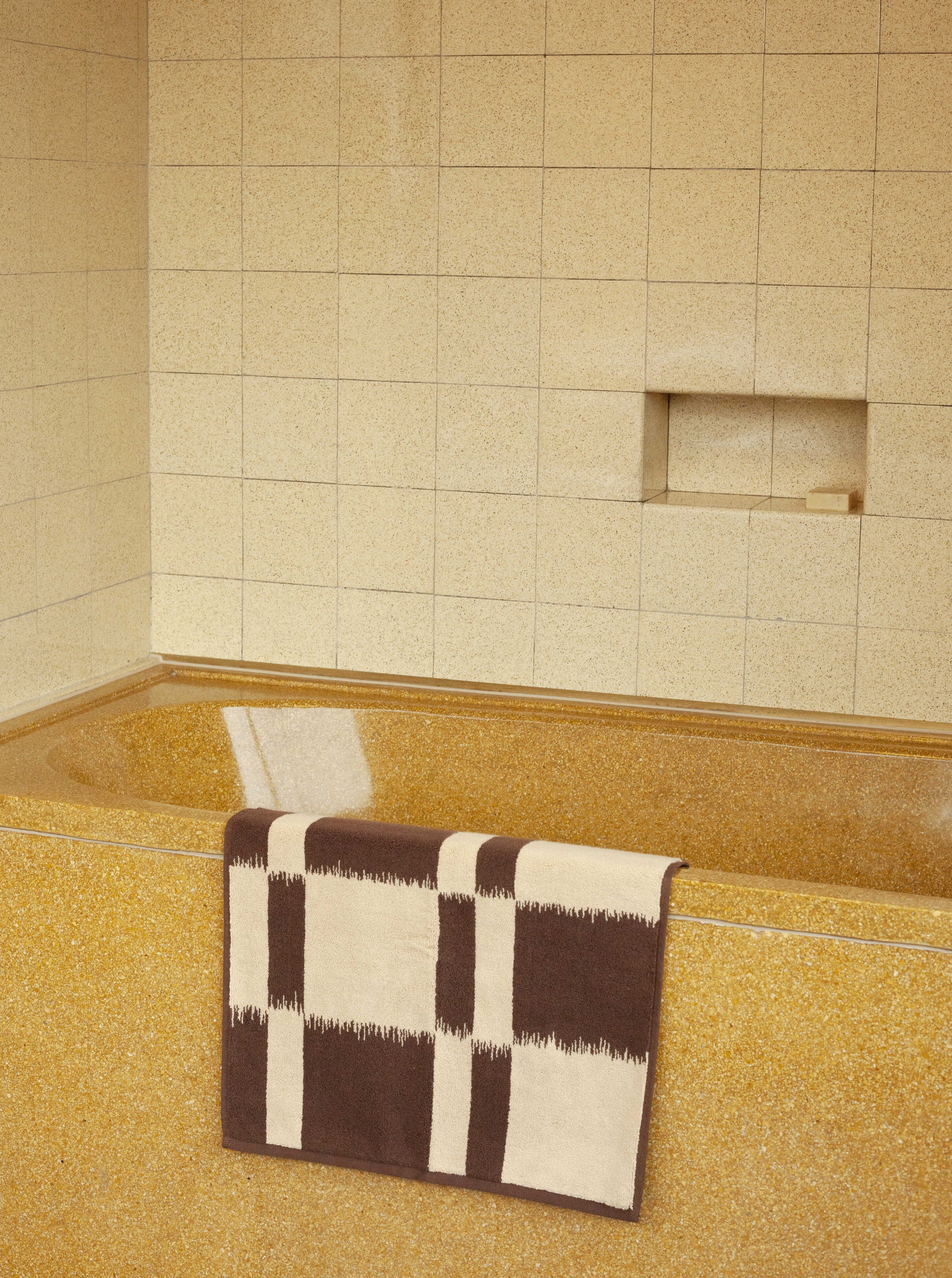 A retro-styled bathroom featuring beige square tiles on the wall, a recessed shelf, and a similar colored speckled floor. An Autumn Sonata Ikat weaving-inspired striped beige and brown towel is draped over a Karin Bath Mat.