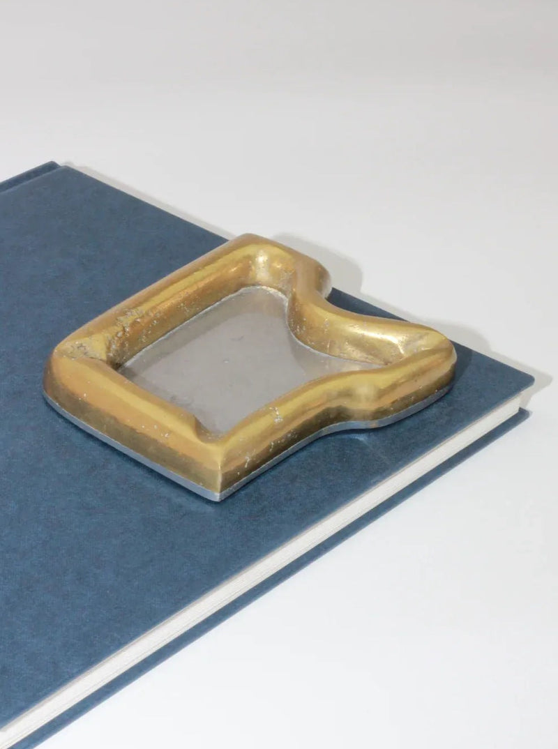 A book with a dark blue cover sits on a white surface. Atop the book rests the Brutalist Ashtray by Boga Avante Shop, with its quirky, rectangular shape featuring a recessed area and worn edges. This vintage item adds an intriguing touch to the scene.