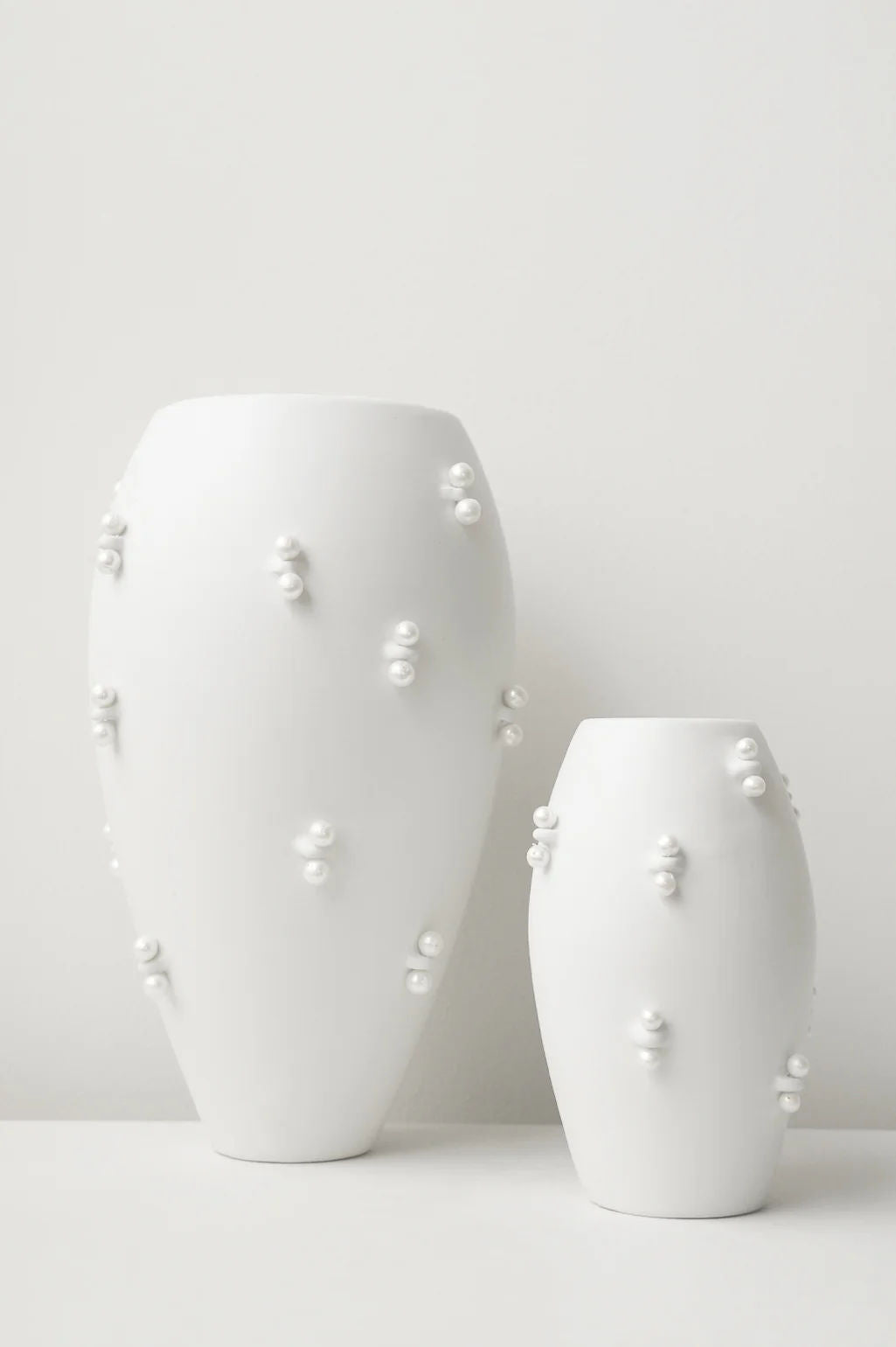 Two "Pinched" vases by Completedworks, one larger and one smaller, feature a matte white ceramic finish with flowing contours and clusters of pearl-like embellishments, set against a plain white background.