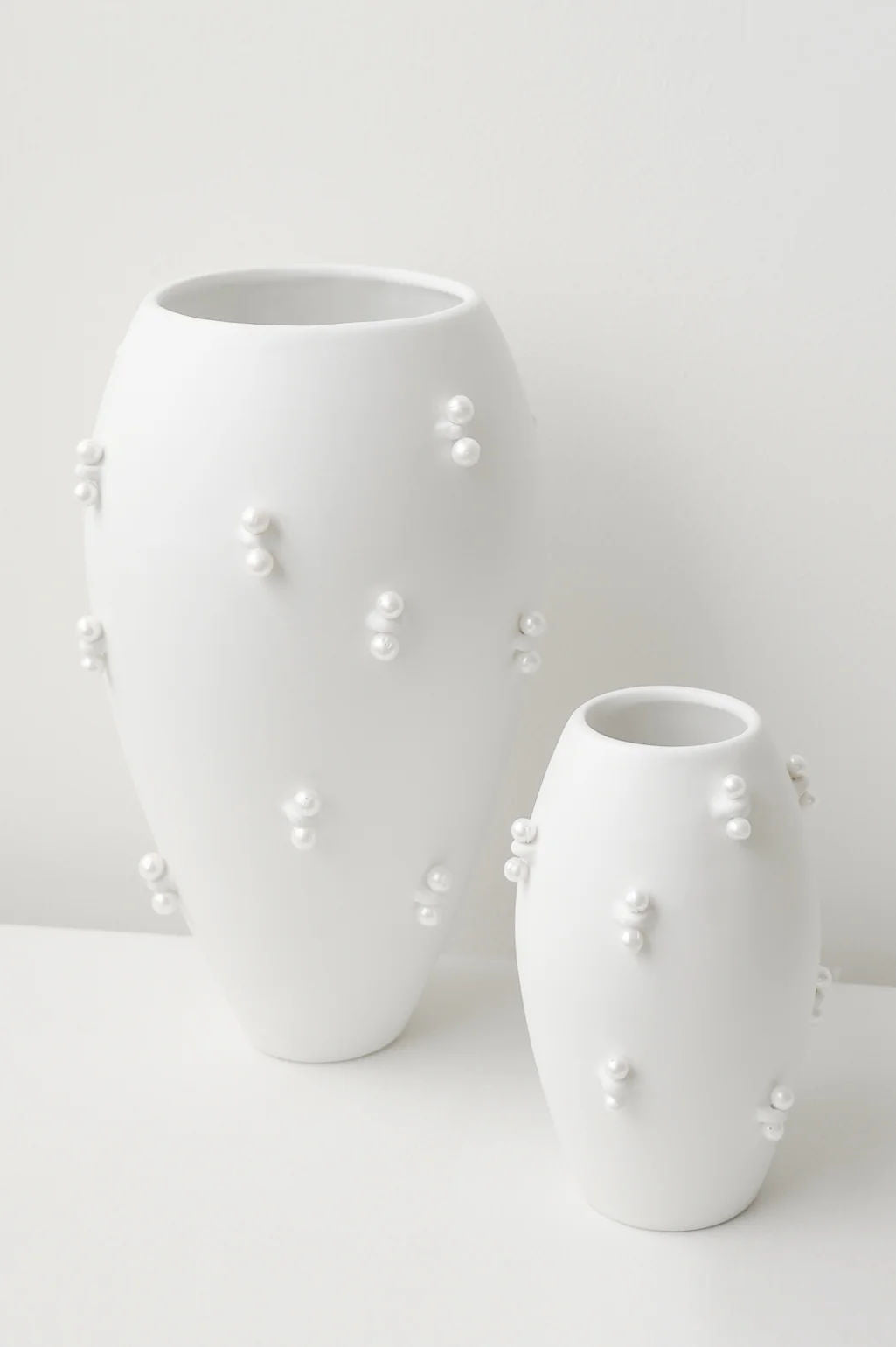 Against a plain backdrop, two vases by Completedworks are featured: the "Pinched" small vase in matte white and its larger counterpart. Both ceramic pieces have smooth, flowing contours adorned with pearl-like embellishments for a textured design.