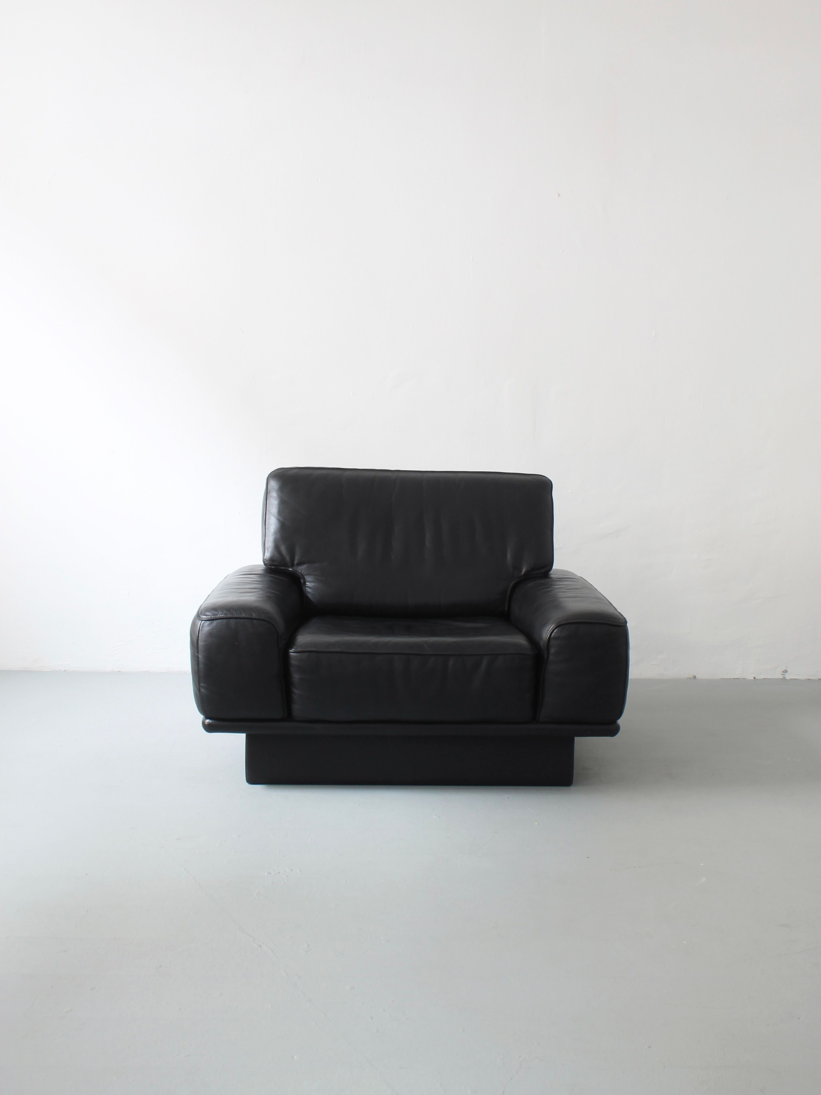 A black leather "Lounge Chair by De Sede" from Afterhour Furniture, showcasing Swiss luxury with cushioned armrests and a sleek, boxy shape, rests on a light gray floor against a plain white wall.