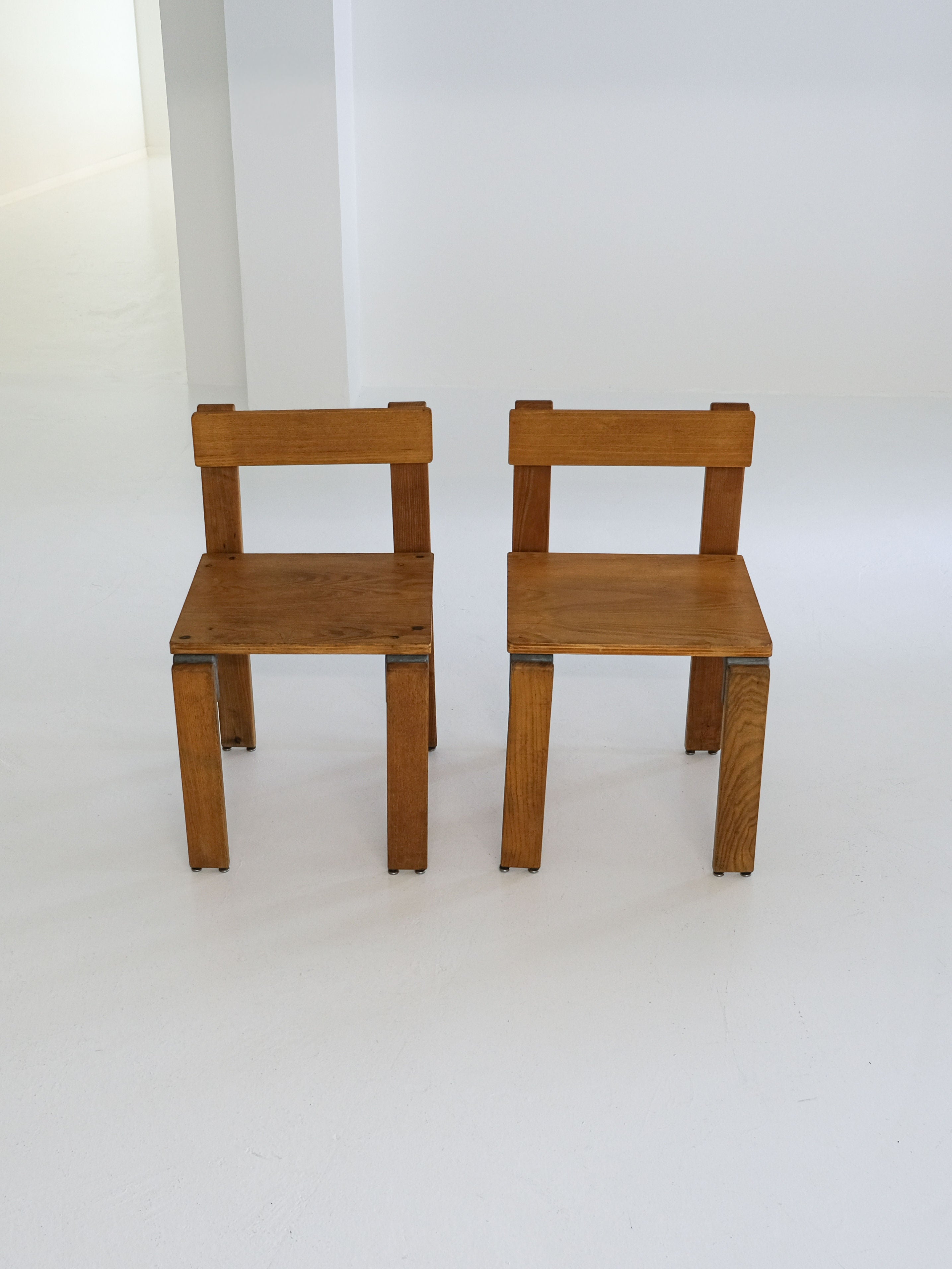 Two "Les Carats" dining chairs by George Candilis and Anja Blomstedt from the 1970s, made by antibeige, are placed side by side on a white floor against a light-colored wall. Crafted from oak plywood, they feature square legs and flat backs, embodying a minimalist vintage style.