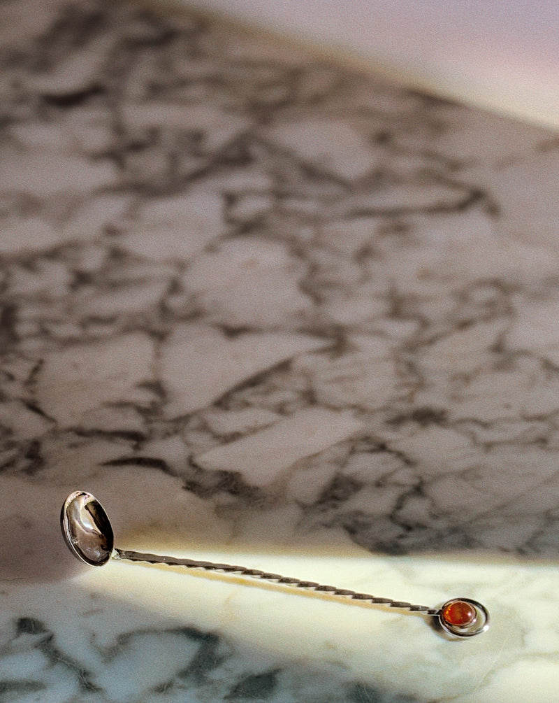 A Huerto Silver Plated Amber Handle Spoon from the set, featuring a twisted handle and amber detail, rests on a sunlit marble surface with gray veining.