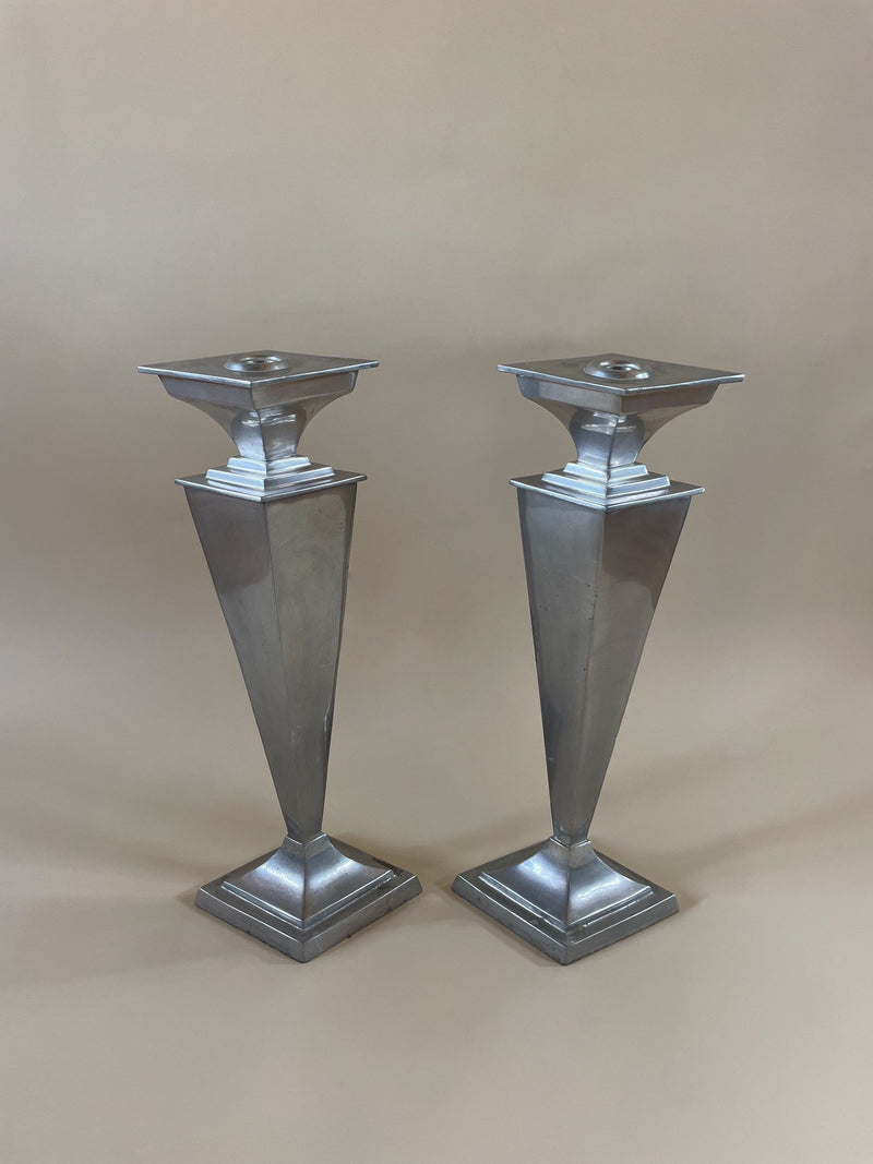The Pair of French Art Deco Candleholders by Maud Vaughan is set against a neutral backdrop, showcasing their silver geometric design. Each piece features a pyramid-like structure with square tops and bases, characterized by angular and symmetrical lines made from metal. These striking candleholders embody a modern, minimalist aesthetic inspired by French Art Deco style.