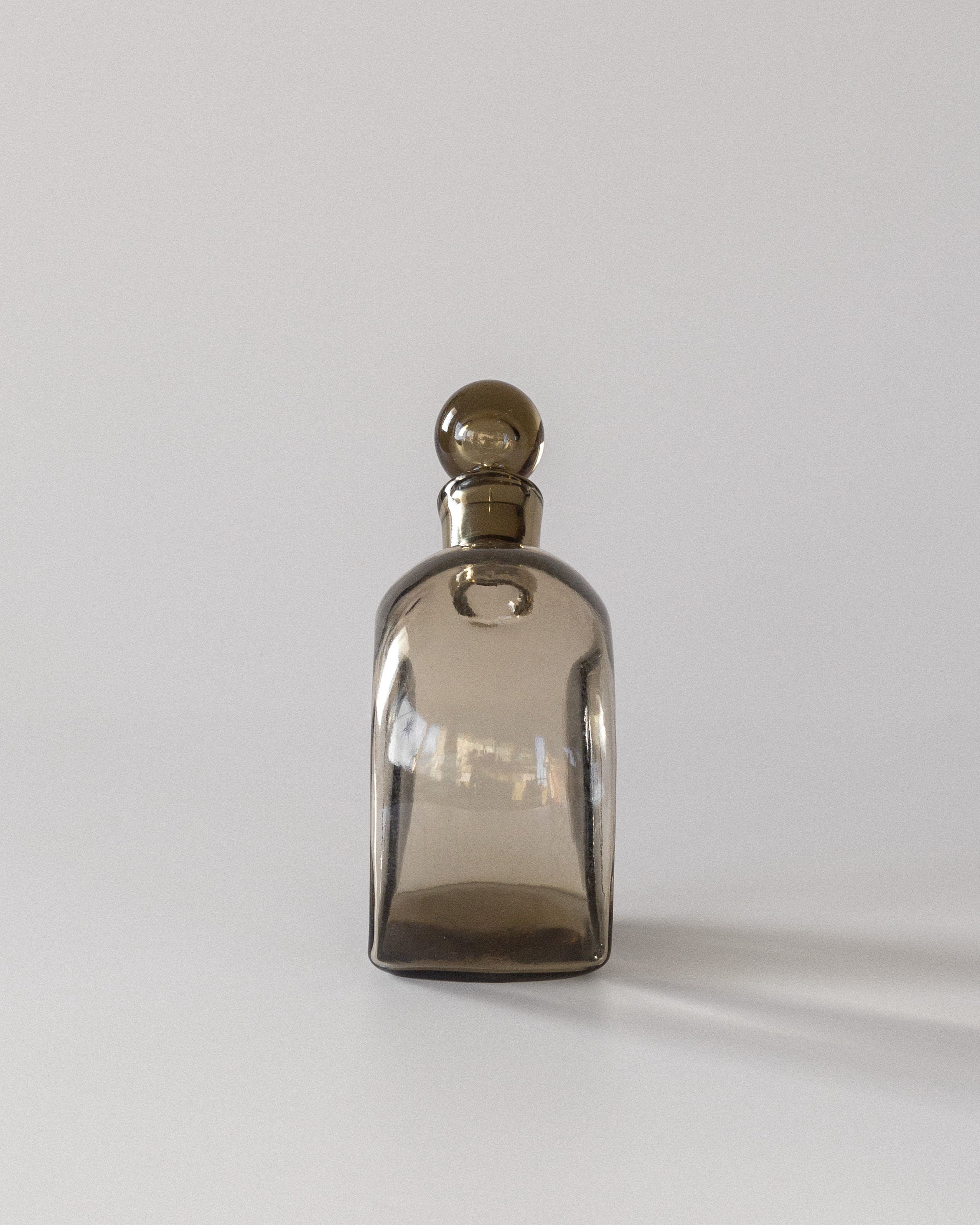 A Glass Decanter by Harald Notini for Spigel, crafted in the 1930s, features elegantly shaped modernist design and a round stopper. Made of smoky brown glass, it casts a shadow on a plain white backdrop, giving it a vintage yet distinctly Swedish modern appeal.