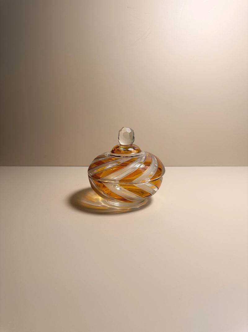 A 1970s Hand-Painted Crystal Bonbonnière from Dodo Vintage, characterized by its round glass design, faceted stopper, and elegant gold and white swirling patterns, rests gracefully on a smooth, reflective surface against a neutral backdrop. Soft illumination heightens its appeal, casting a gentle shadow.