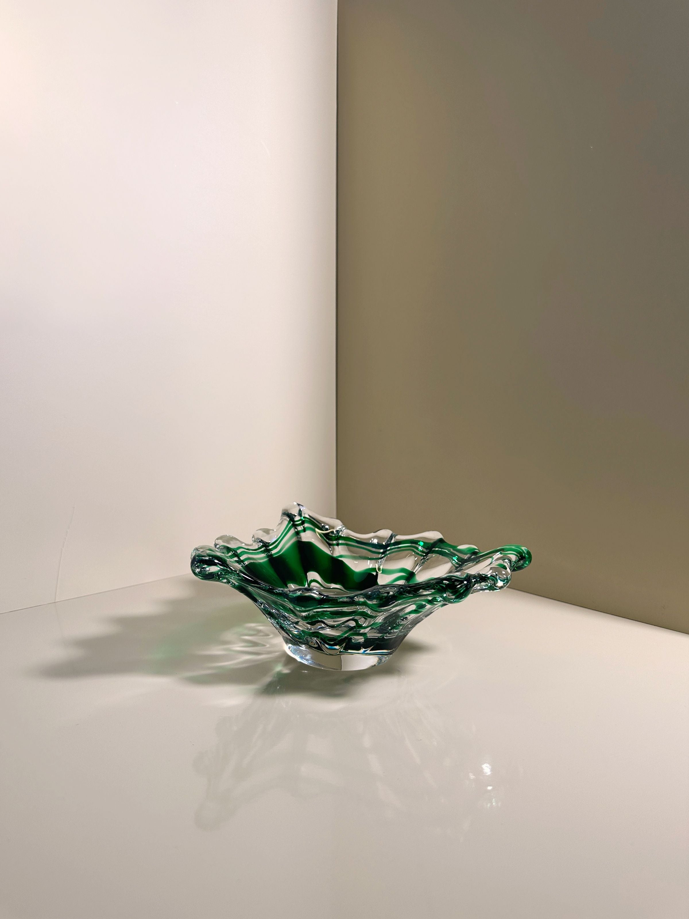 A Green Swirled Murano Vide-Poche by Dodo Vintage sits on a reflective white surface, its wavy edges casting shadows that highlight its intricate design. The background showcases intersecting white and beige walls.