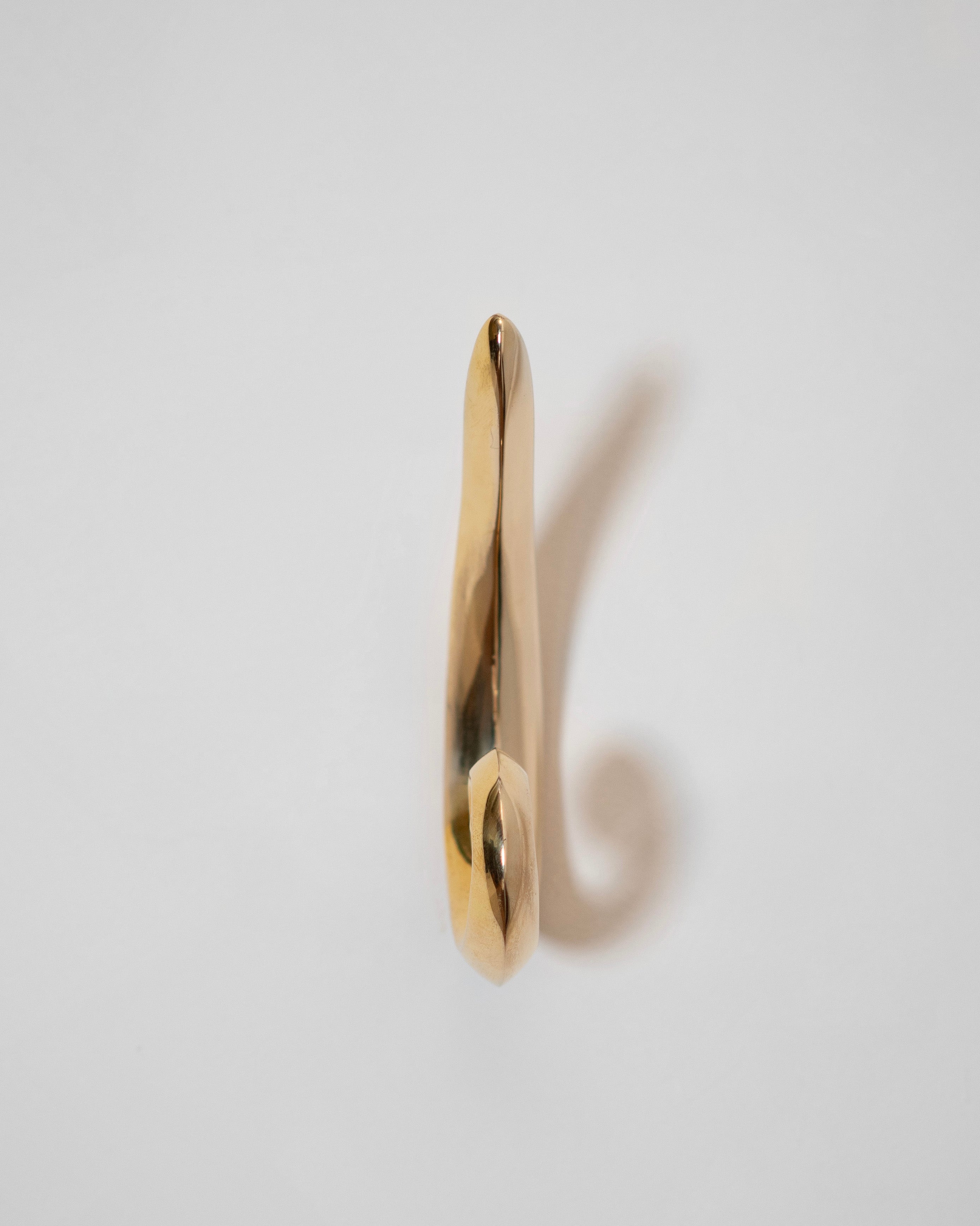 The Noelle Hook - Brass by Hein Studio is a single, shiny brass sandcast hook mounted on a white surface, viewed from the front, casting a soft shadow to the right.