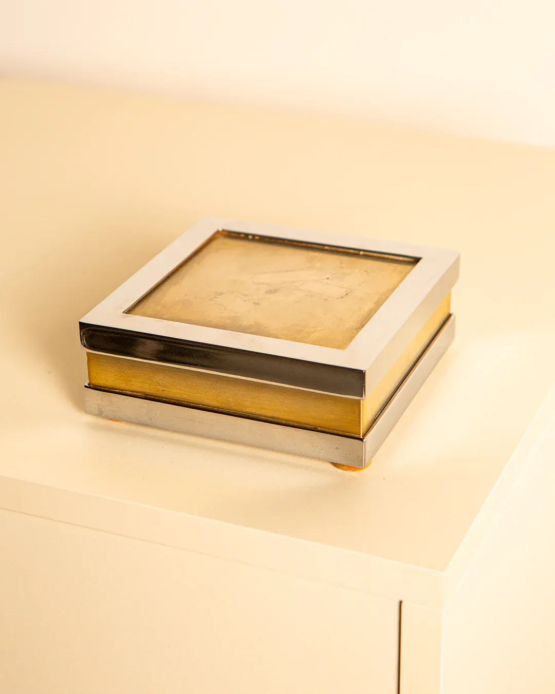 The Treaptyque Italian Silver-plated Metal and Brass Box 60's, a small square with a glass top, rests on a light surface. Its brass band elegantly enhances the reflective silver-plated frame, harmonizing beautifully with the neutral-toned background.