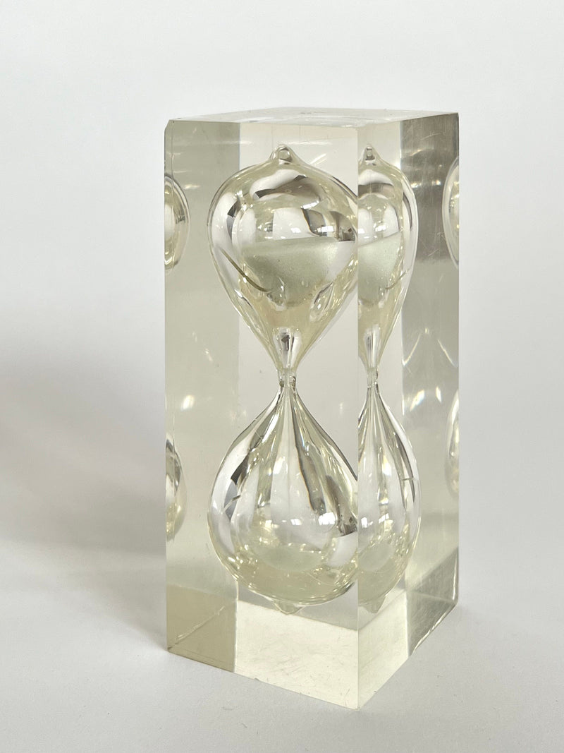 A clear, rectangular lucite cube encases the "Hourglass by Charles Hollis Jones, 1970s" from septembre studios, creating an illusion of multiple reflections. The hourglass appears suspended at the center, framed by smooth, transparent surfaces reminiscent of Jones’s innovative designs.